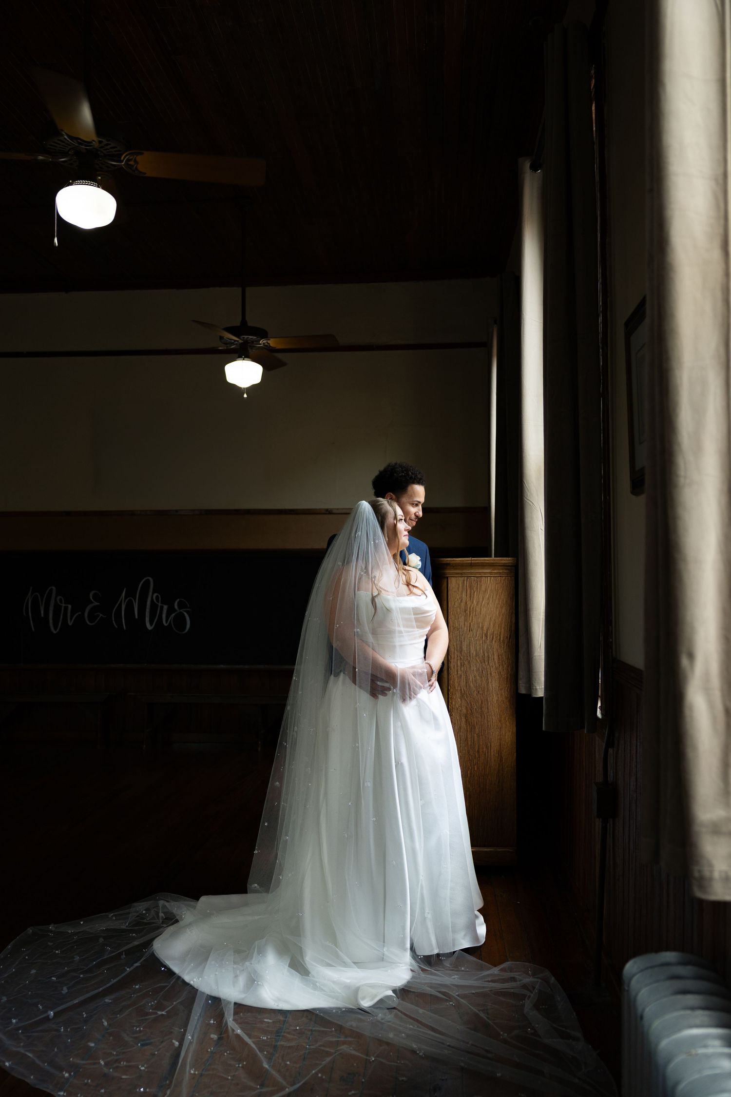 Bride and groom embracing in soft window light inside rustic church venue.