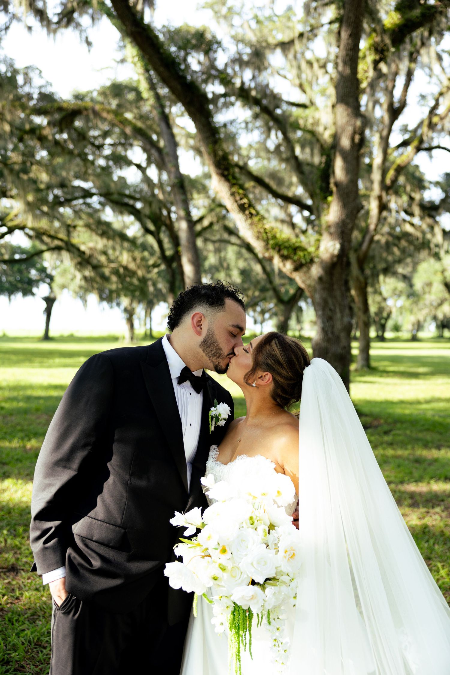 Bride and groom share a romantic kiss under moss-draped oak trees, bride holding white orchid bouquet in elegant wedding attire.