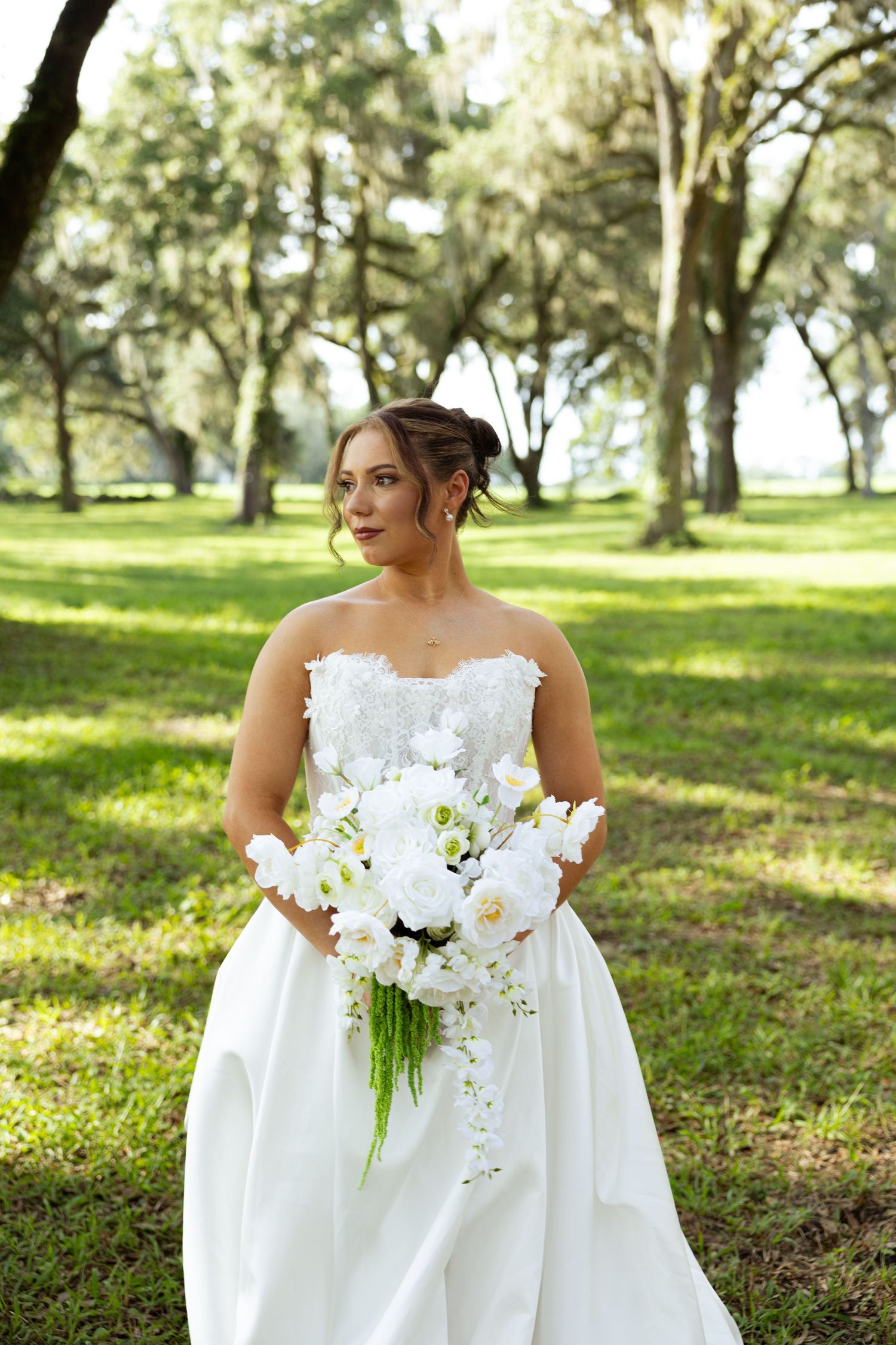 Bride in strapless white gown holding white floral bouquet outdoors in lush green park setting.