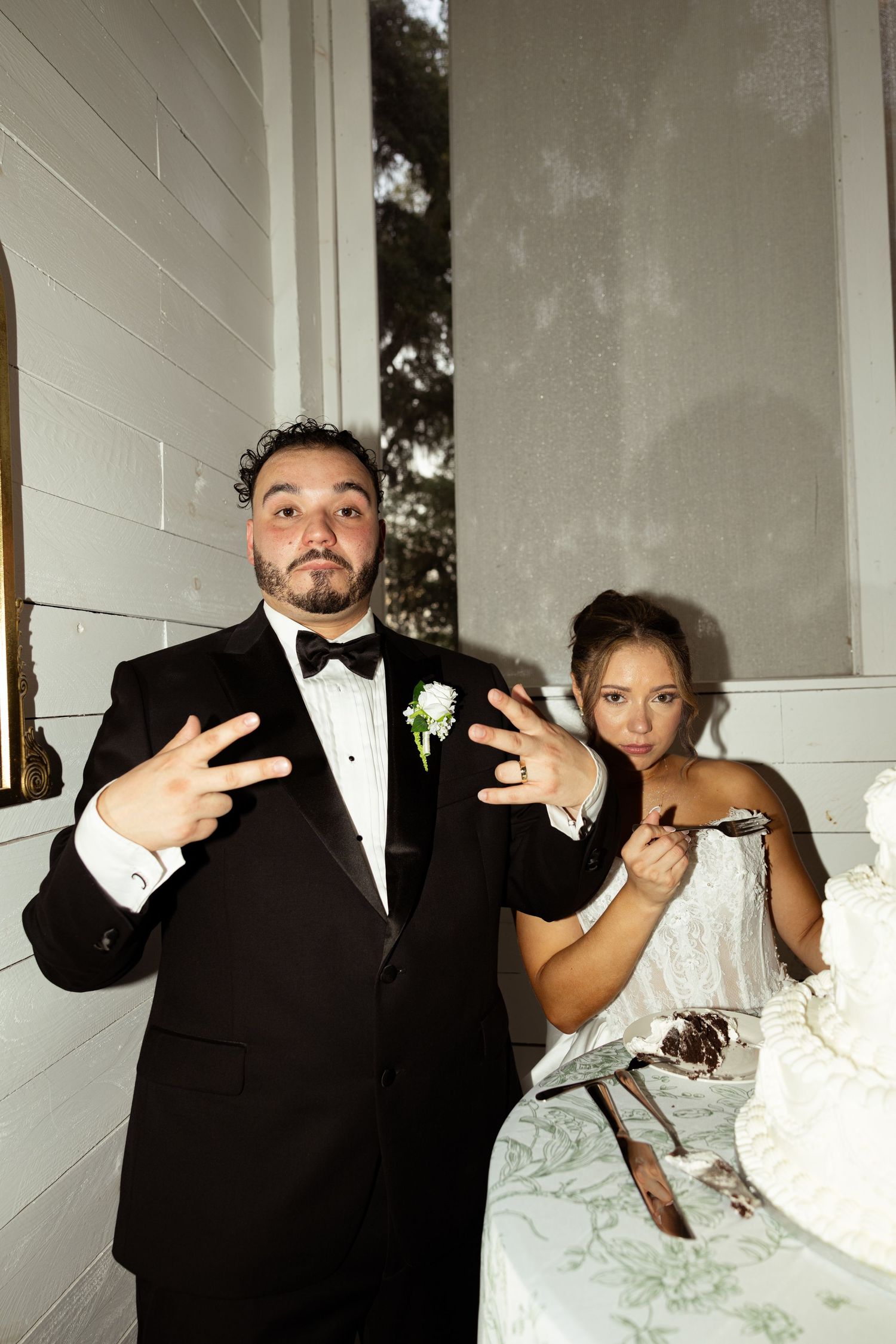 Groom in tuxedo posing with peace signs while bride cuts wedding cake at elegant reception.
