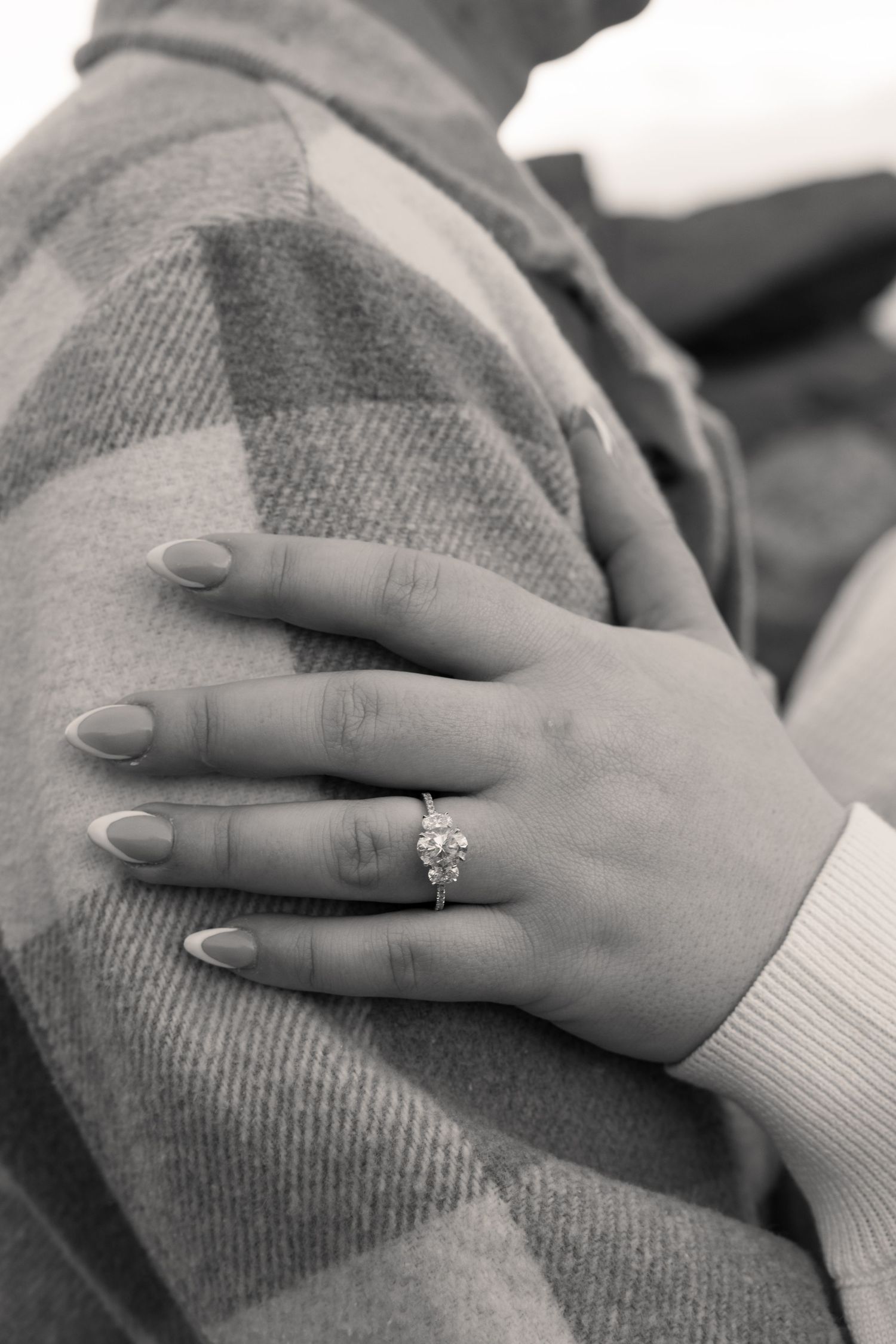 Close-up black and white photo of a womans hand with engagement ring resting on a mans plaid jacket shoulder.