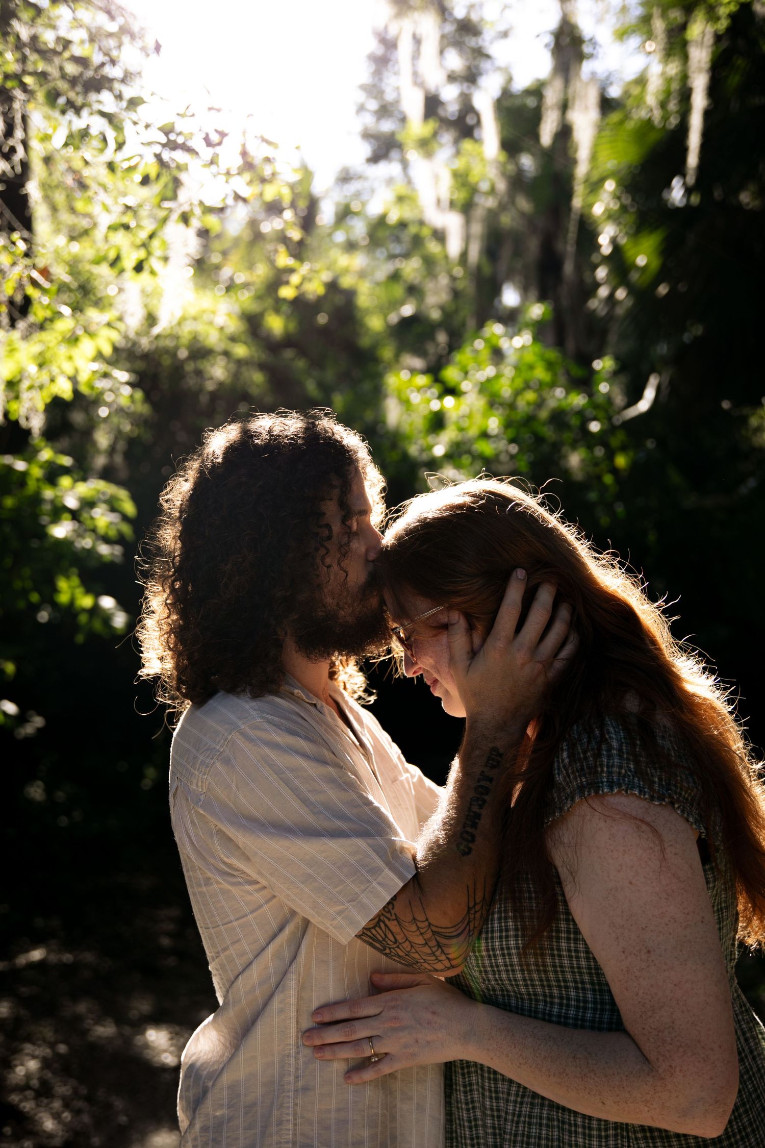 A couple shares a tender moment in a sunlit forest, the man gently cradling the woman's face as they lean in close.