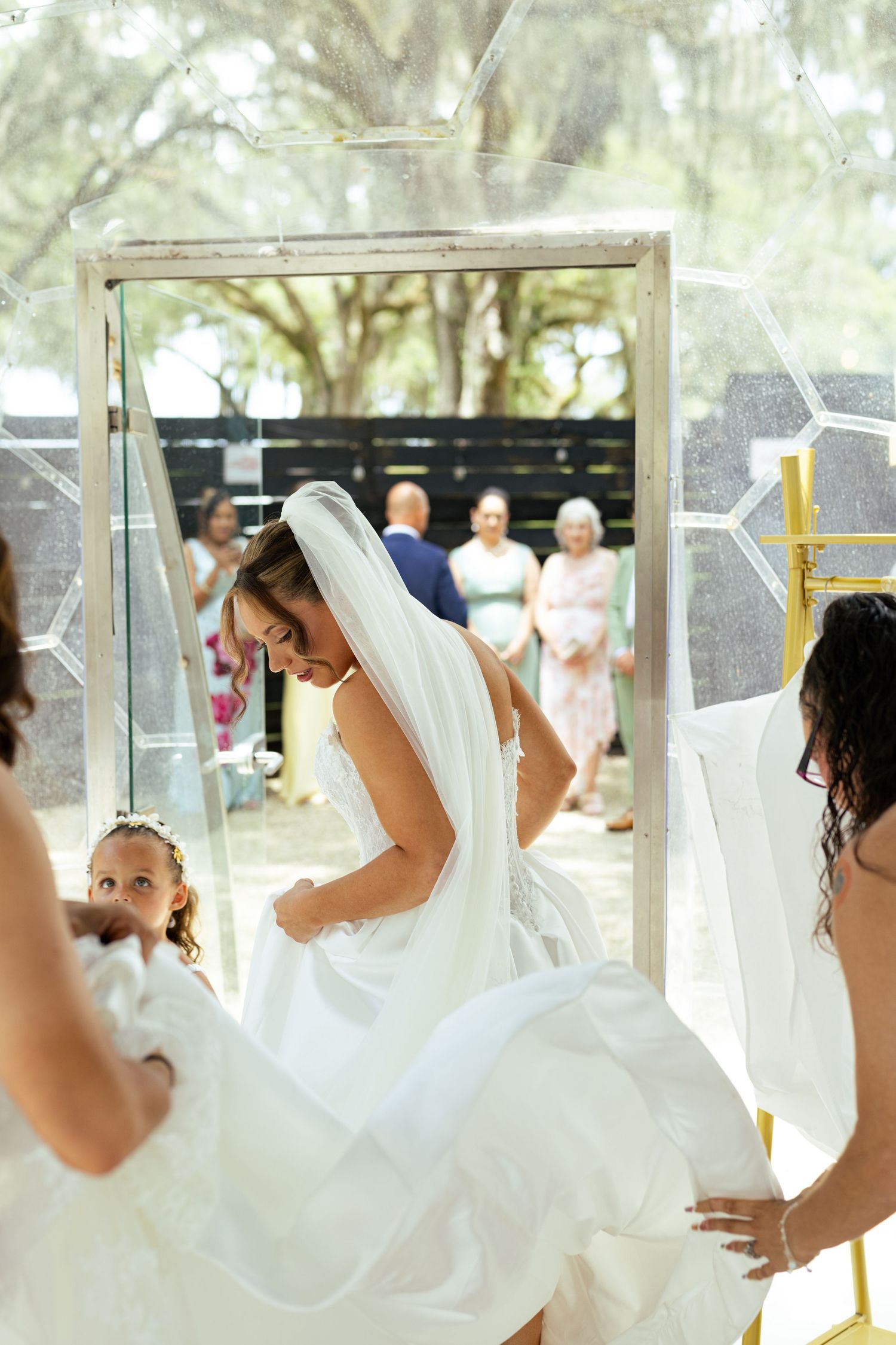 Bride in white gown and veil adjusting her dress with bridesmaids help at an outdoor wedding ceremony.