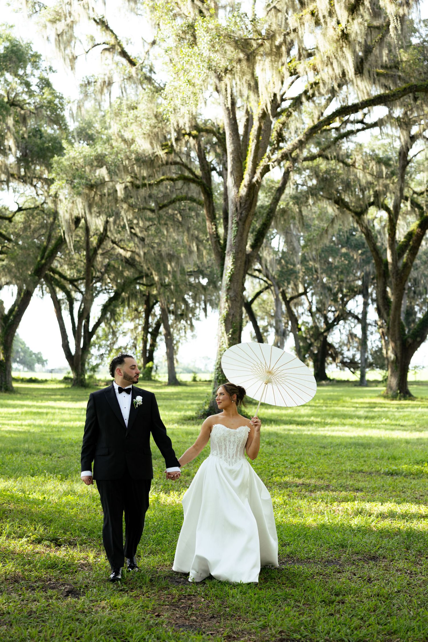 Bride and groom walk hand-in-hand through a sunlit park with Spanish moss trees, bride holding a white parasol.