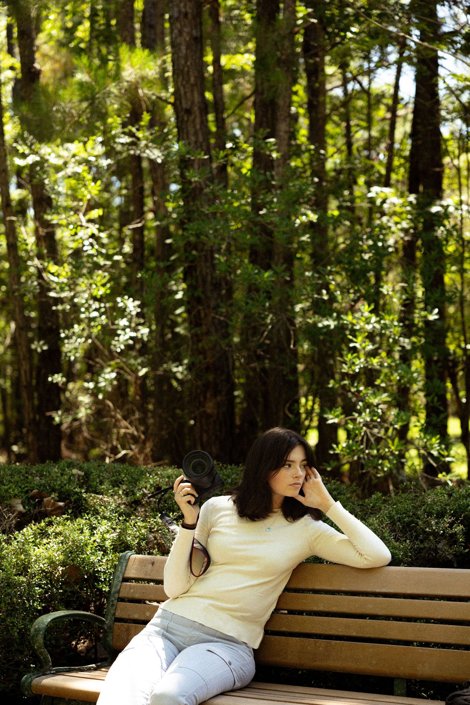 Young woman in cream sweater relaxing on wooden park bench surrounded by lush green forest trees.