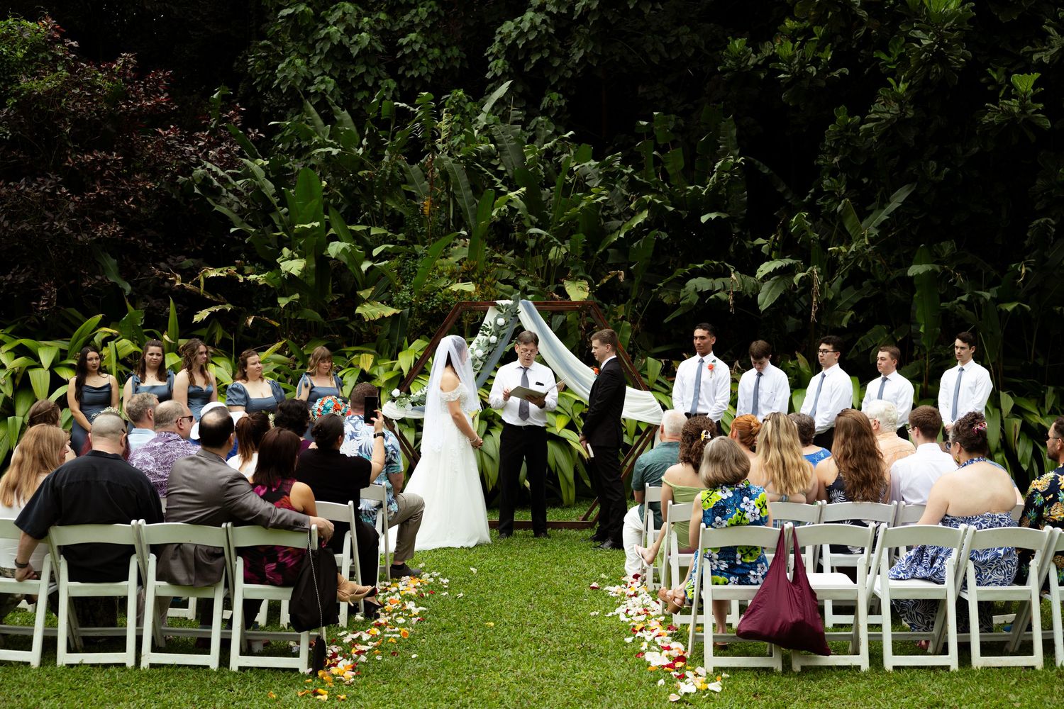 Outdoor wedding ceremony on green lawn with bride and groom at altar, guests seated in white chairs, lush tropical foliage backdrop.