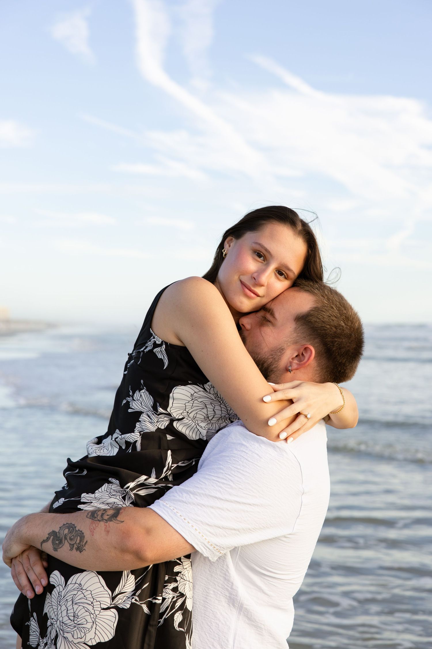 A couple embraces romantically on a beach, the man lifting the woman in a black floral dress against a blue sky backdrop.