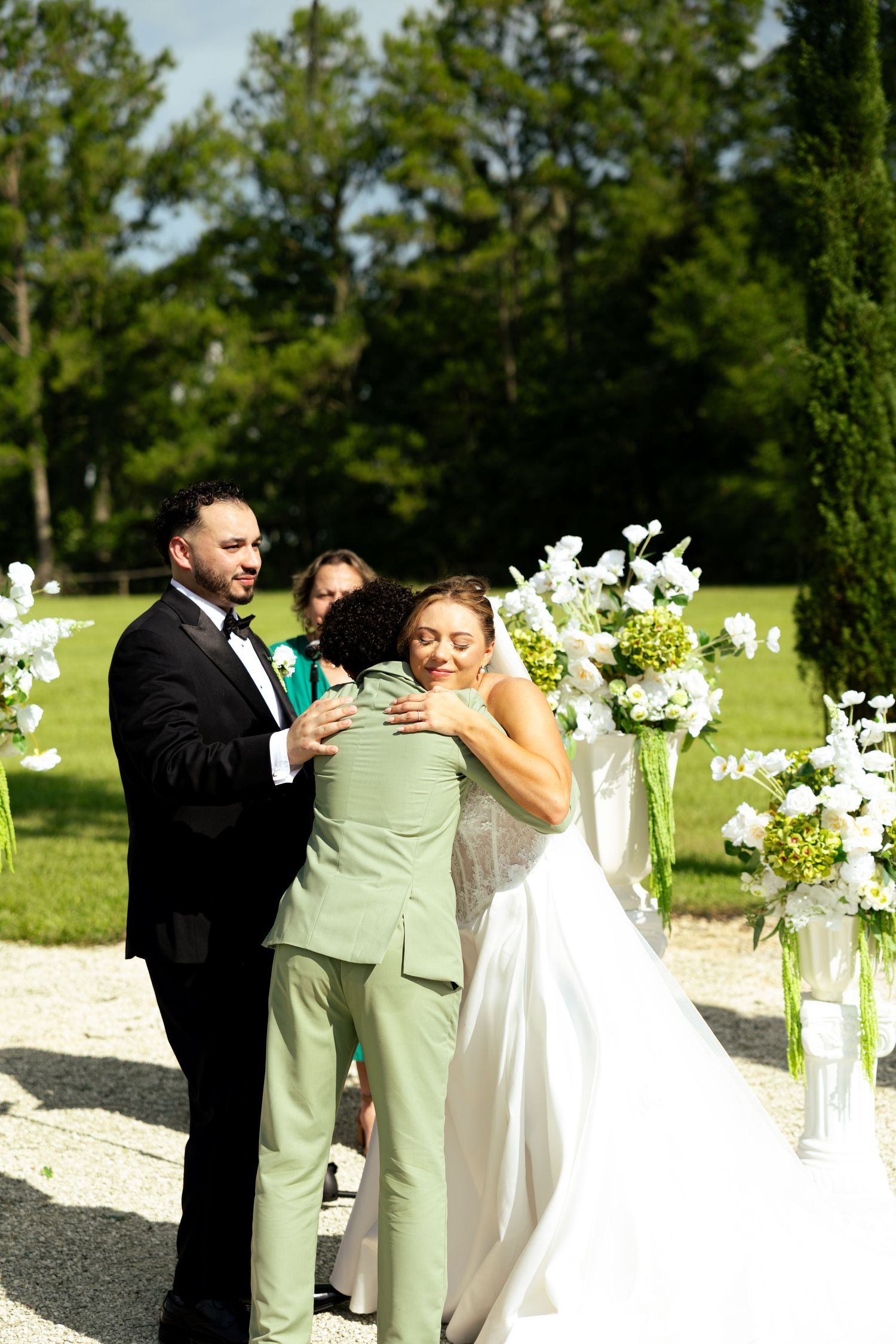 Bride and groom embrace guest at outdoor wedding altar adorned with white floral arrangements.