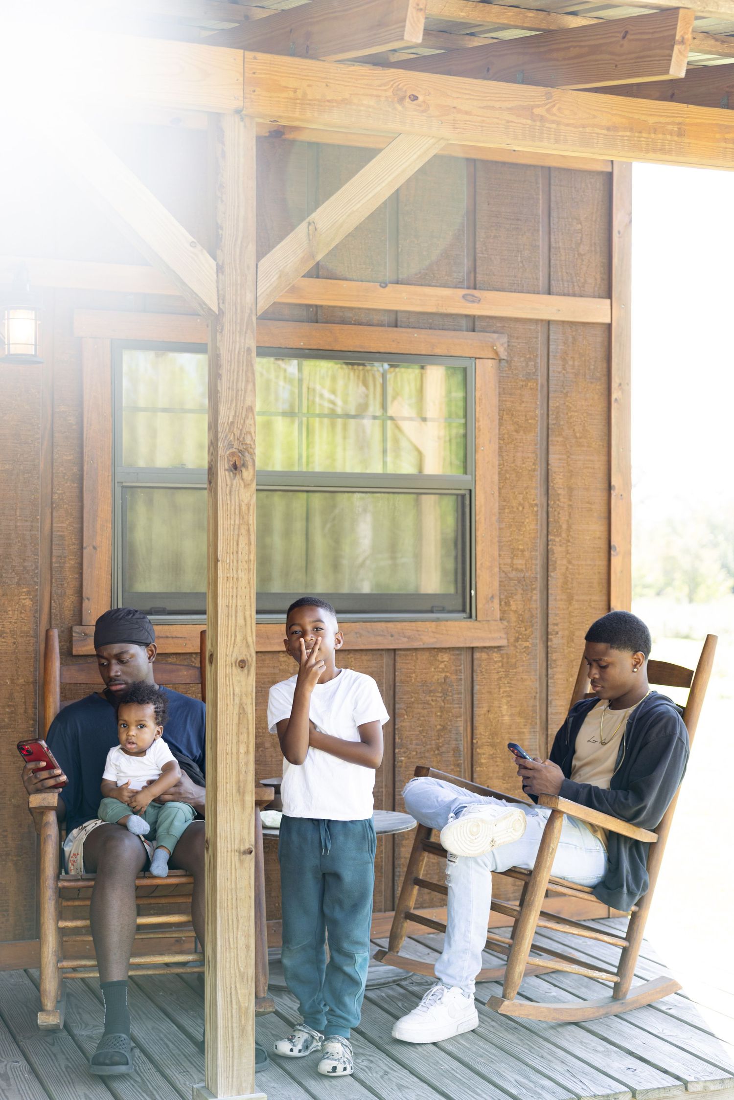 Three young Black men relax on a wooden cabin porch, two sitting in rocking chairs with phones while a child sits on one mans lap.