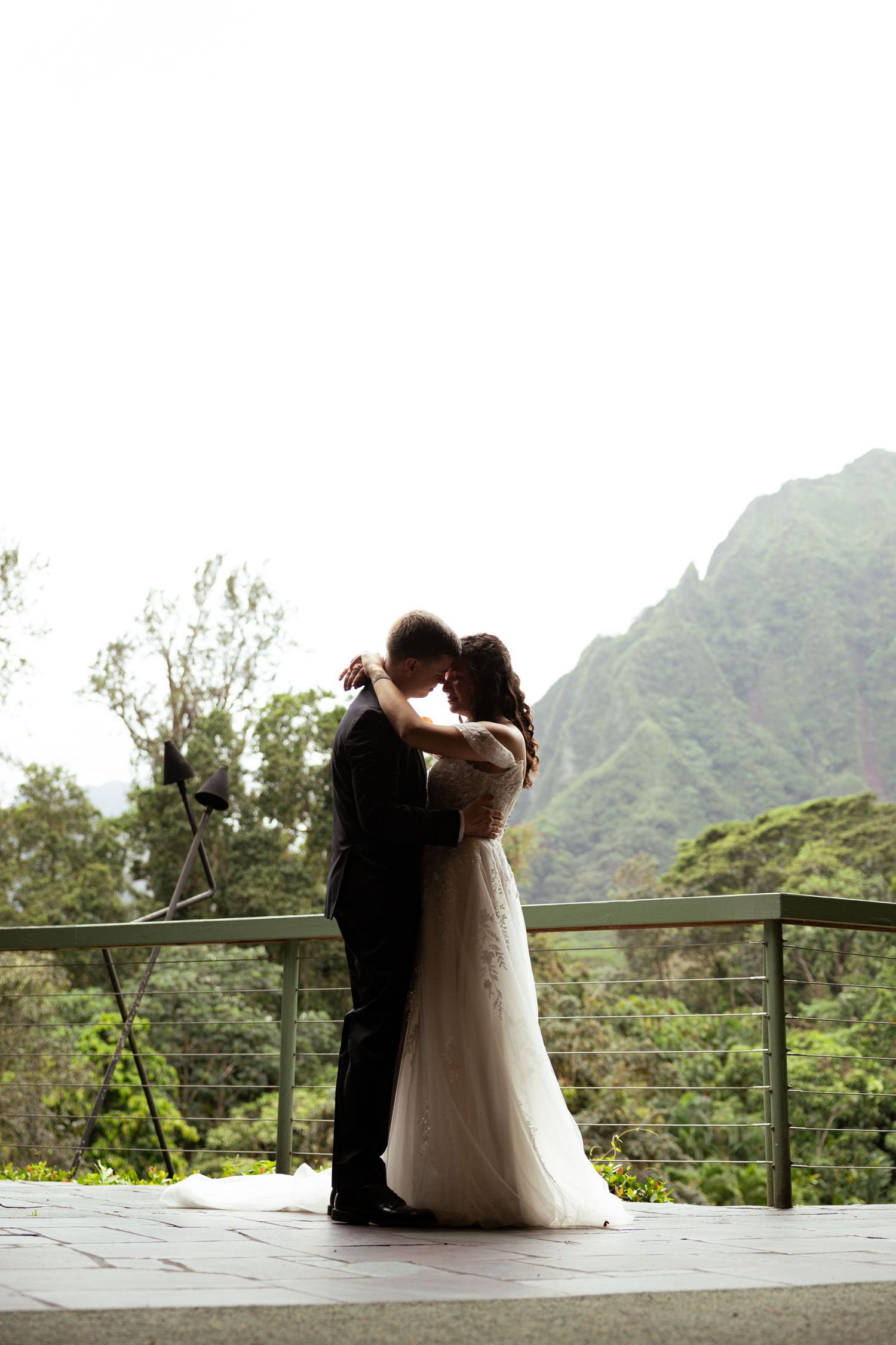 Bride and groom share a romantic kiss on a scenic outdoor terrace with lush green mountains in the background.