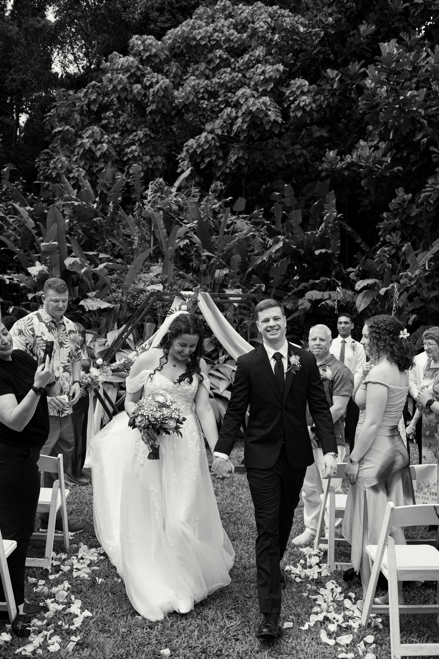 Black and white photo of a smiling bride and groom walking down the aisle after their outdoor wedding ceremony.