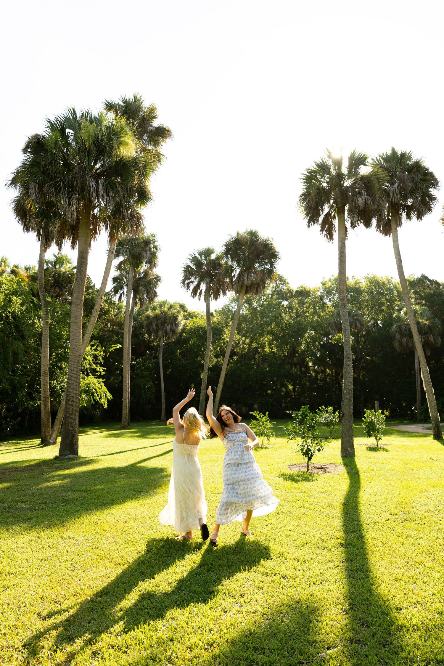 Two women in floral dresses dancing joyfully on green lawn surrounded by tall palm trees in sunny tropical setting.