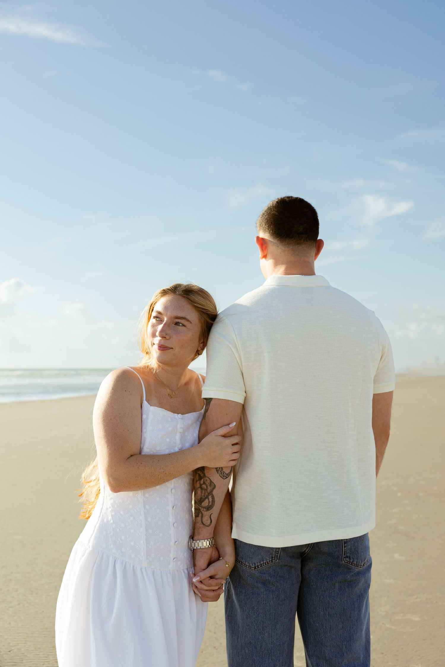 A couple holds hands on a sandy beach, woman in white dress leaning on mans arm under a blue sky.