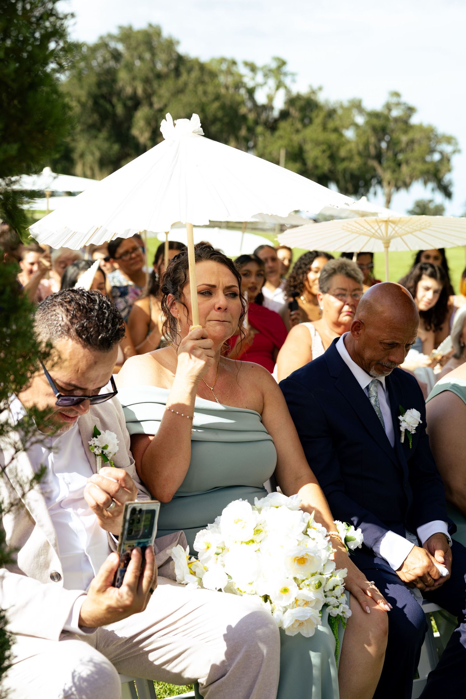 mom Emotional wedding guests watch outdoor ceremony, woman in sage bridesmaid dress holds white bouquet.