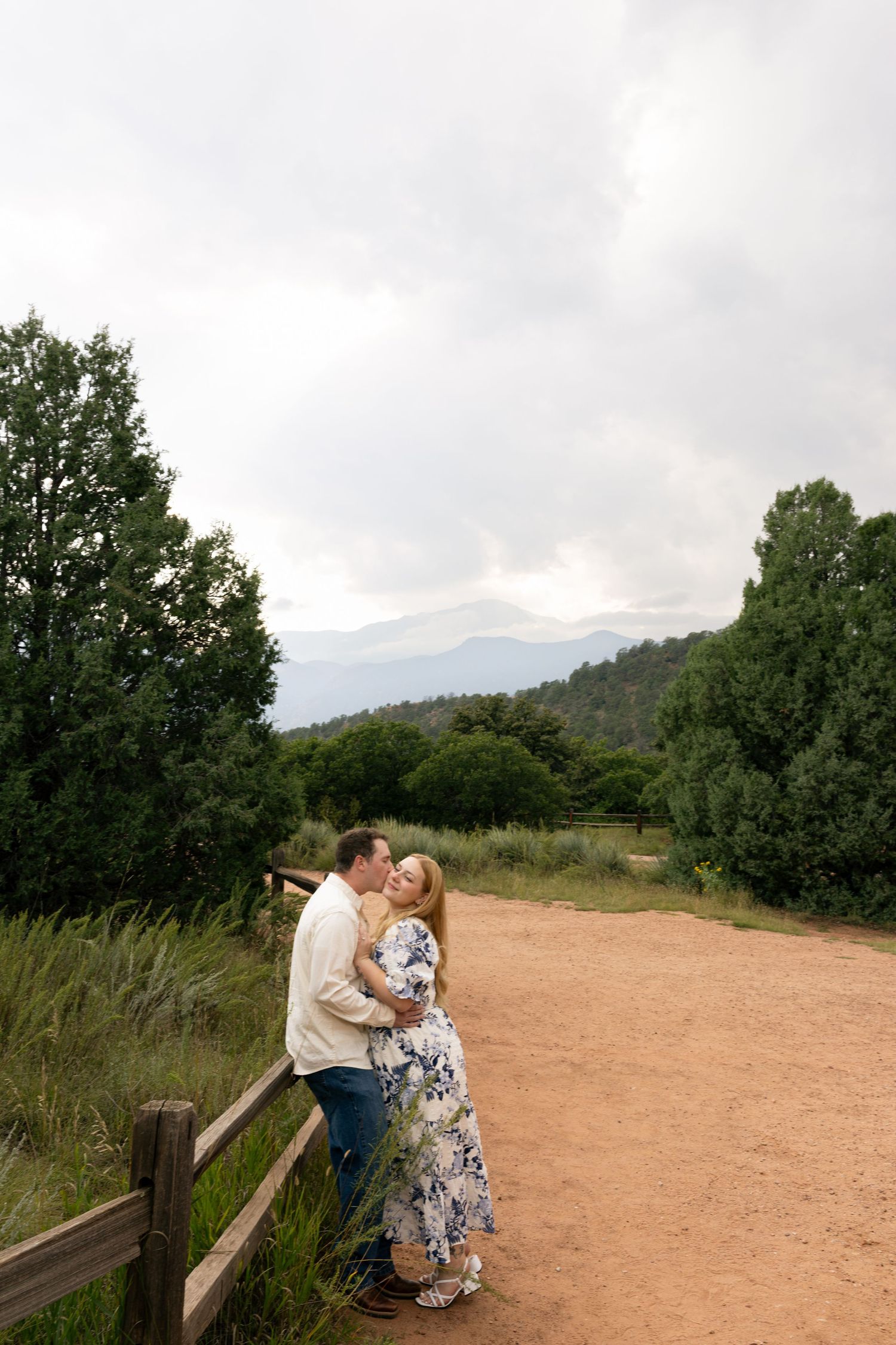 Couple sharing a romantic embrace on a dirt path surrounded by lush green trees and mountains under cloudy skies.