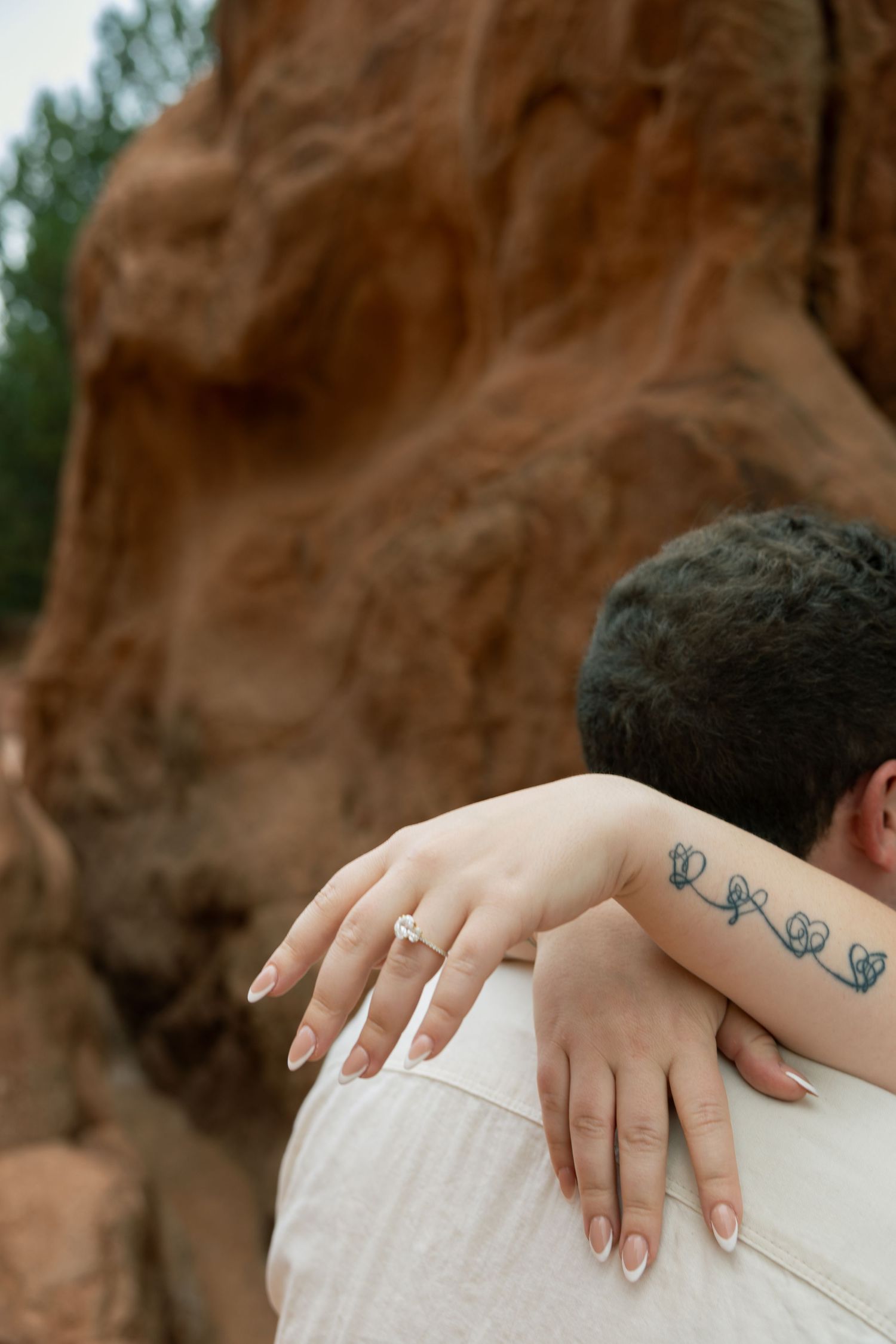 Close-up of woman's hand with diamond engagement ring and floral tattoo resting on man's shoulder at Garden of the Gods.