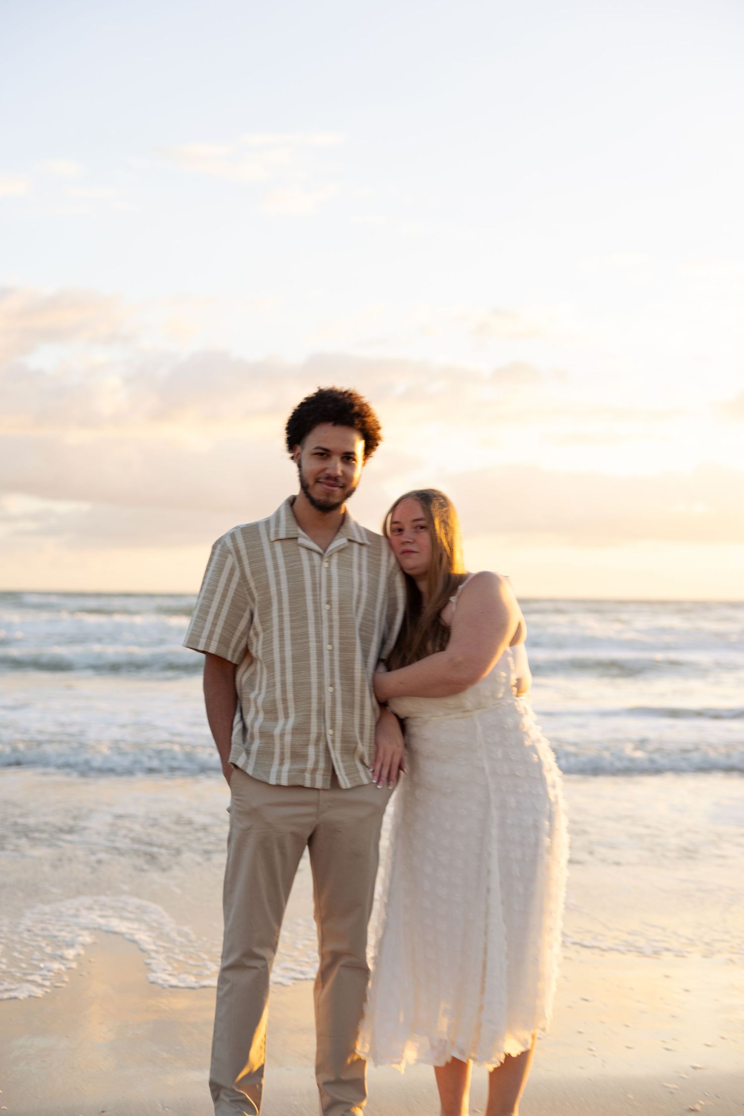 Couple posing together on a sandy beach at sunset, man in striped shirt and woman in white dress.