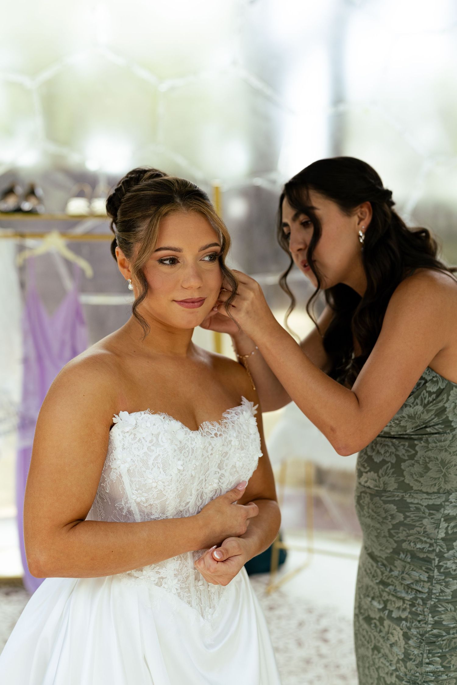 Bridesmaid helps bride with earring while bride wears strapless lace wedding gown before ceremony.