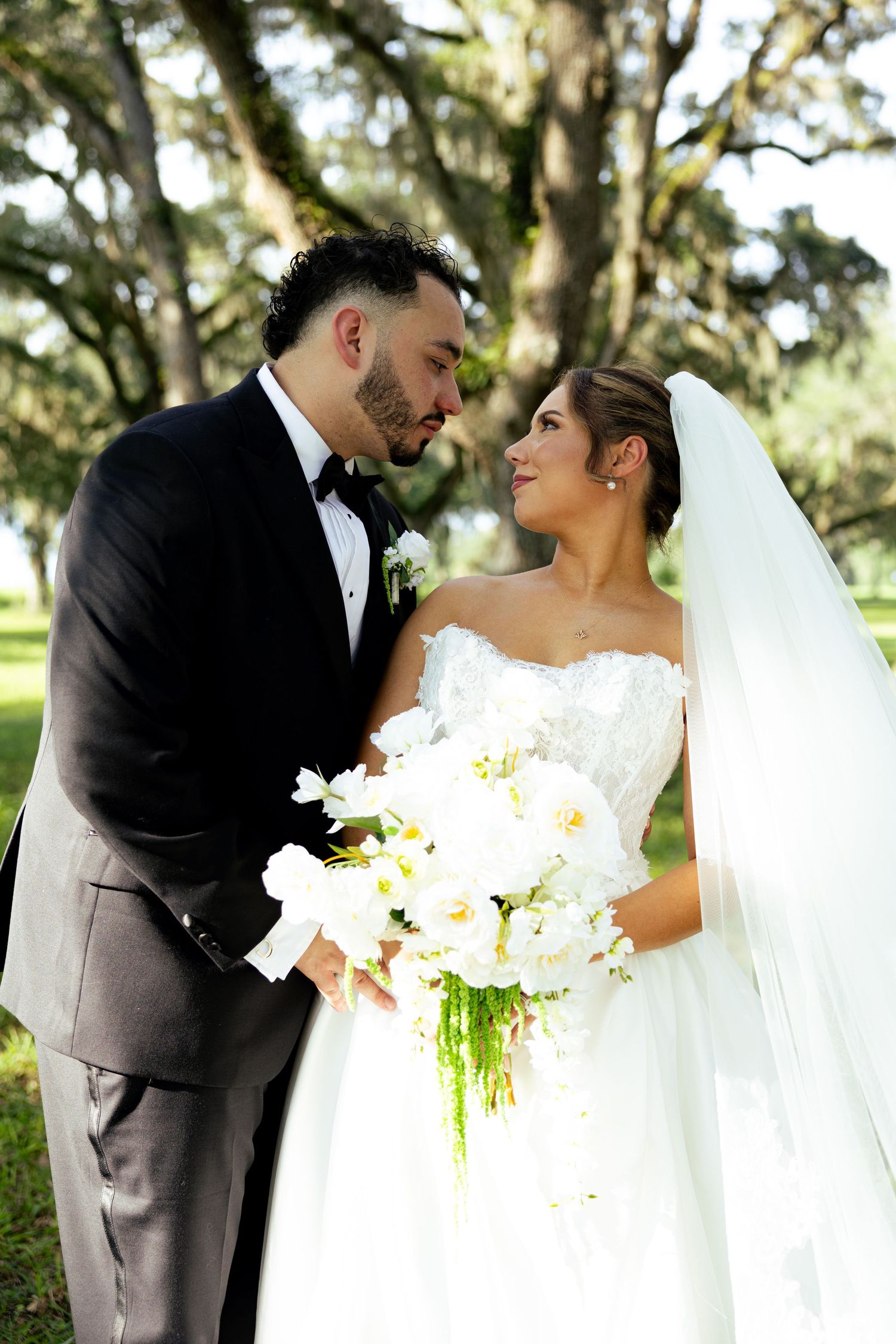 Bride and groom share a romantic moment outdoors, she holds white bouquet, wearing gown and veil, he in black tuxedo.