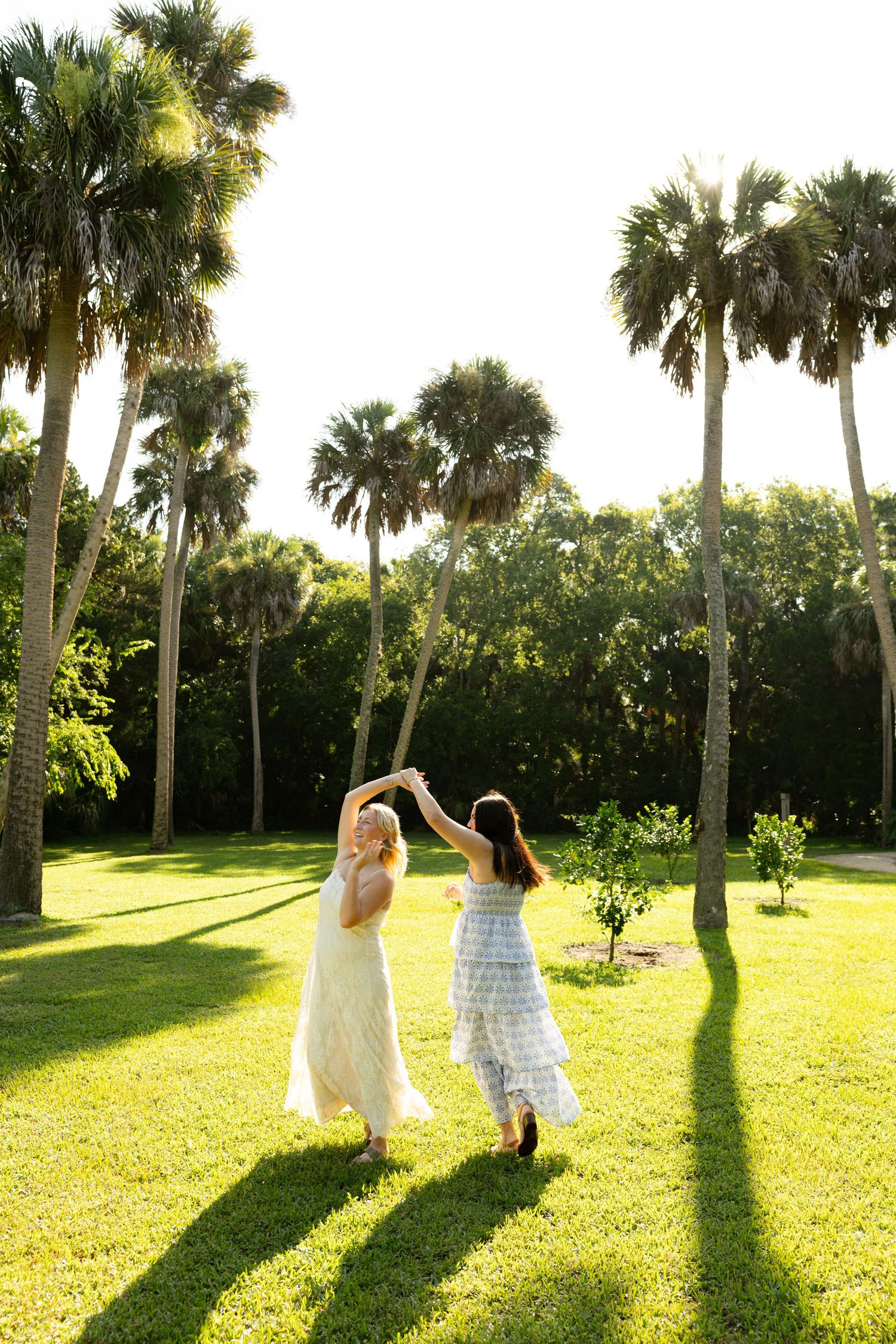 Two women in flowy dresses dancing together on lush green grass surrounded by tall palm trees in golden sunlight.