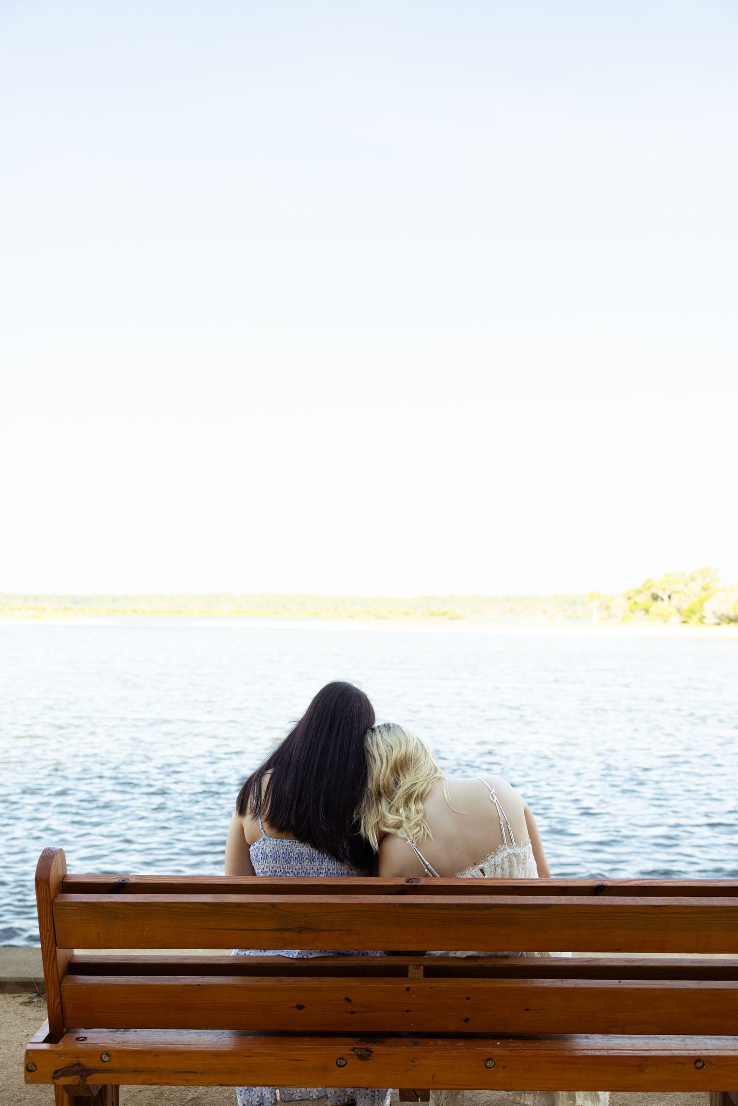 Two women sitting together on a wooden bench overlooking a calm lake, viewed from behind.