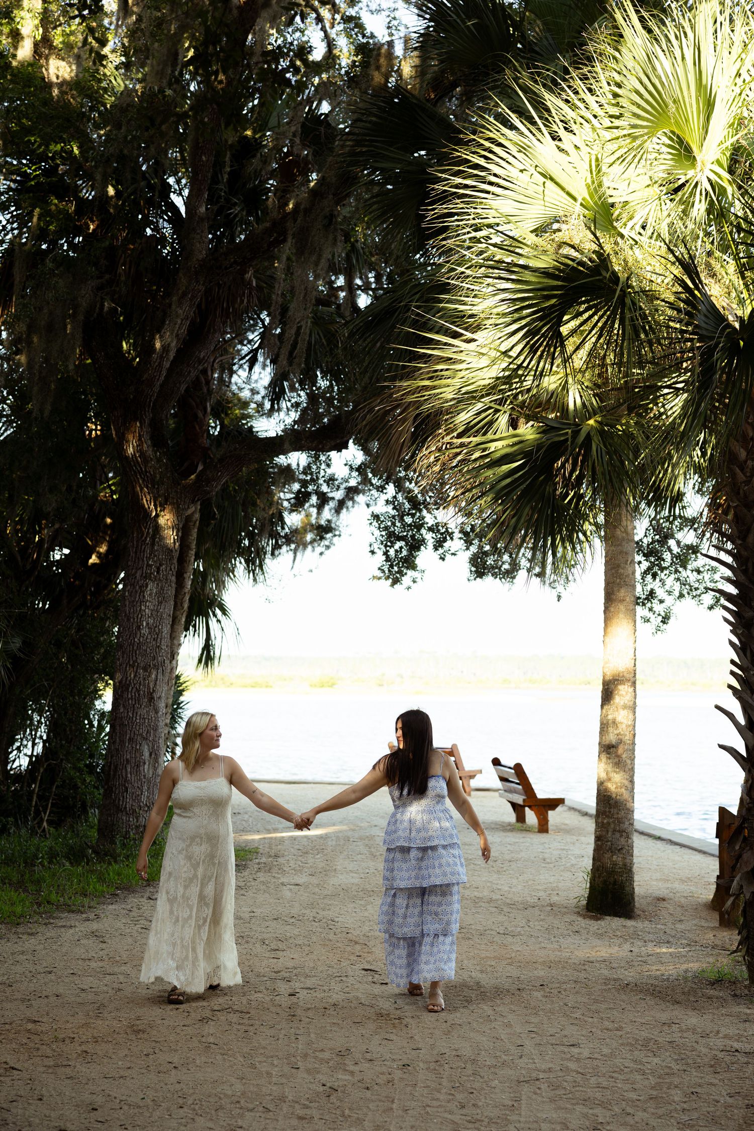 Two women in elegant dresses hold hands while walking along a sandy path lined with palm trees near a sunlit waterfront.