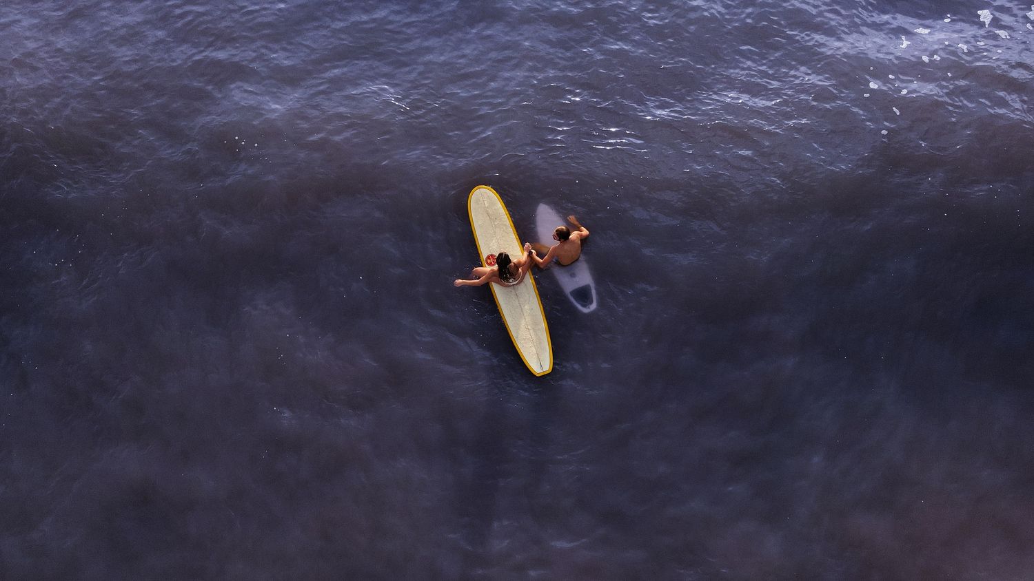 Aerial view of two surfers lying on yellow and black surfboards floating on dark ocean water.