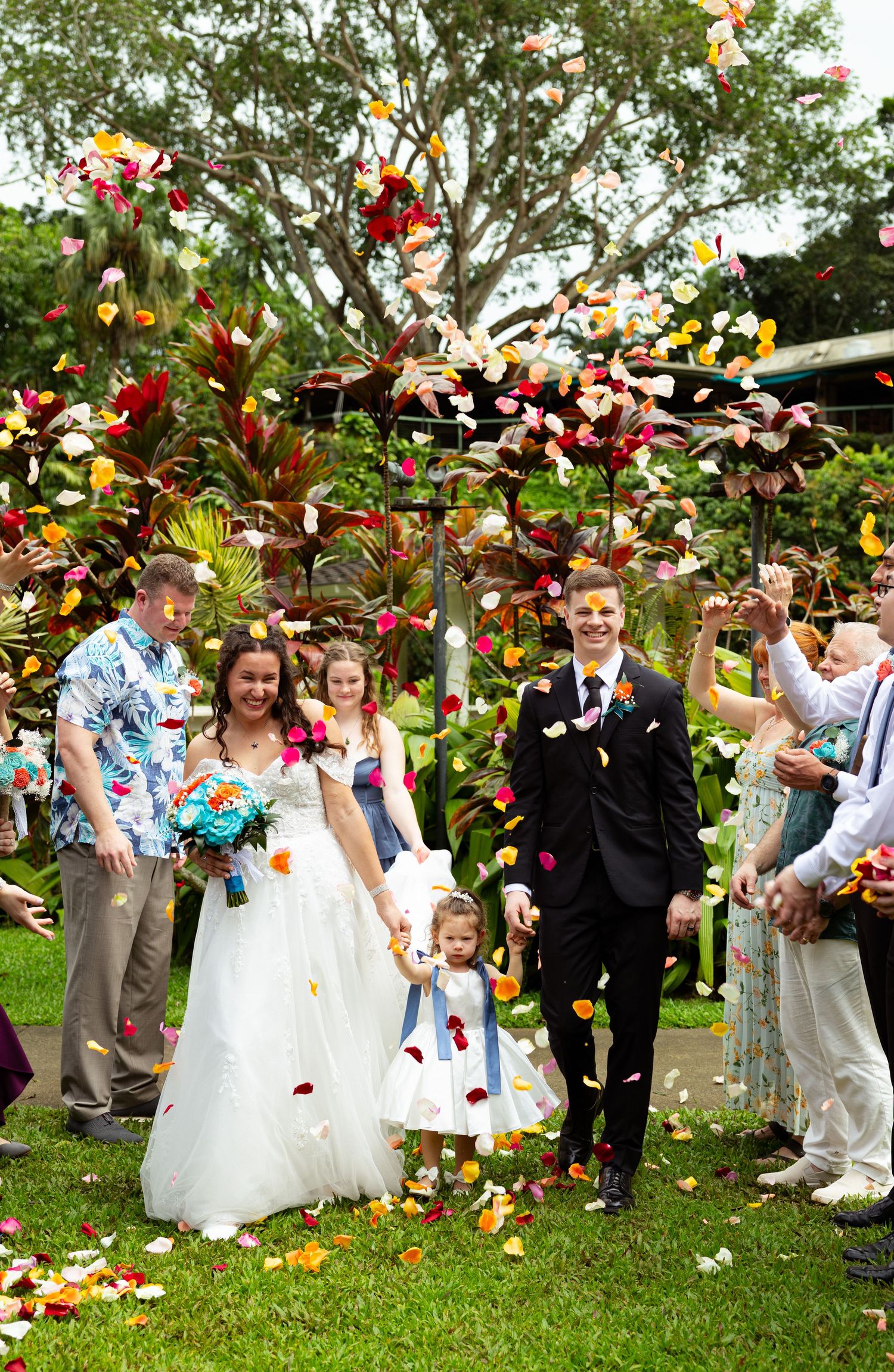 Bride and groom walk down the aisle as guests toss colorful flower petals at an outdoor tropical wedding ceremony.