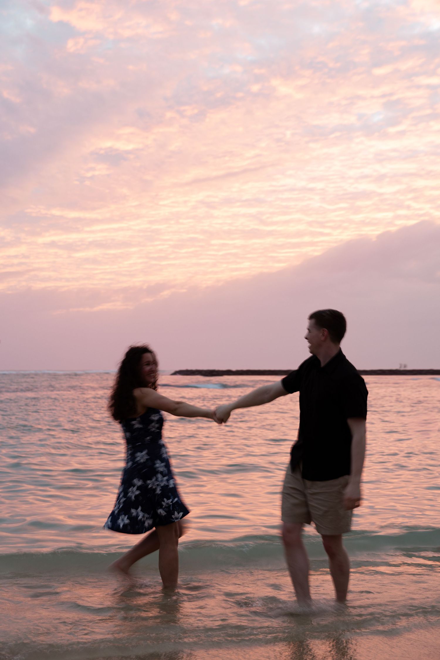 Couple holding hands and dancing in shallow ocean water at a beautiful pink and purple sunset on a tropical beach.