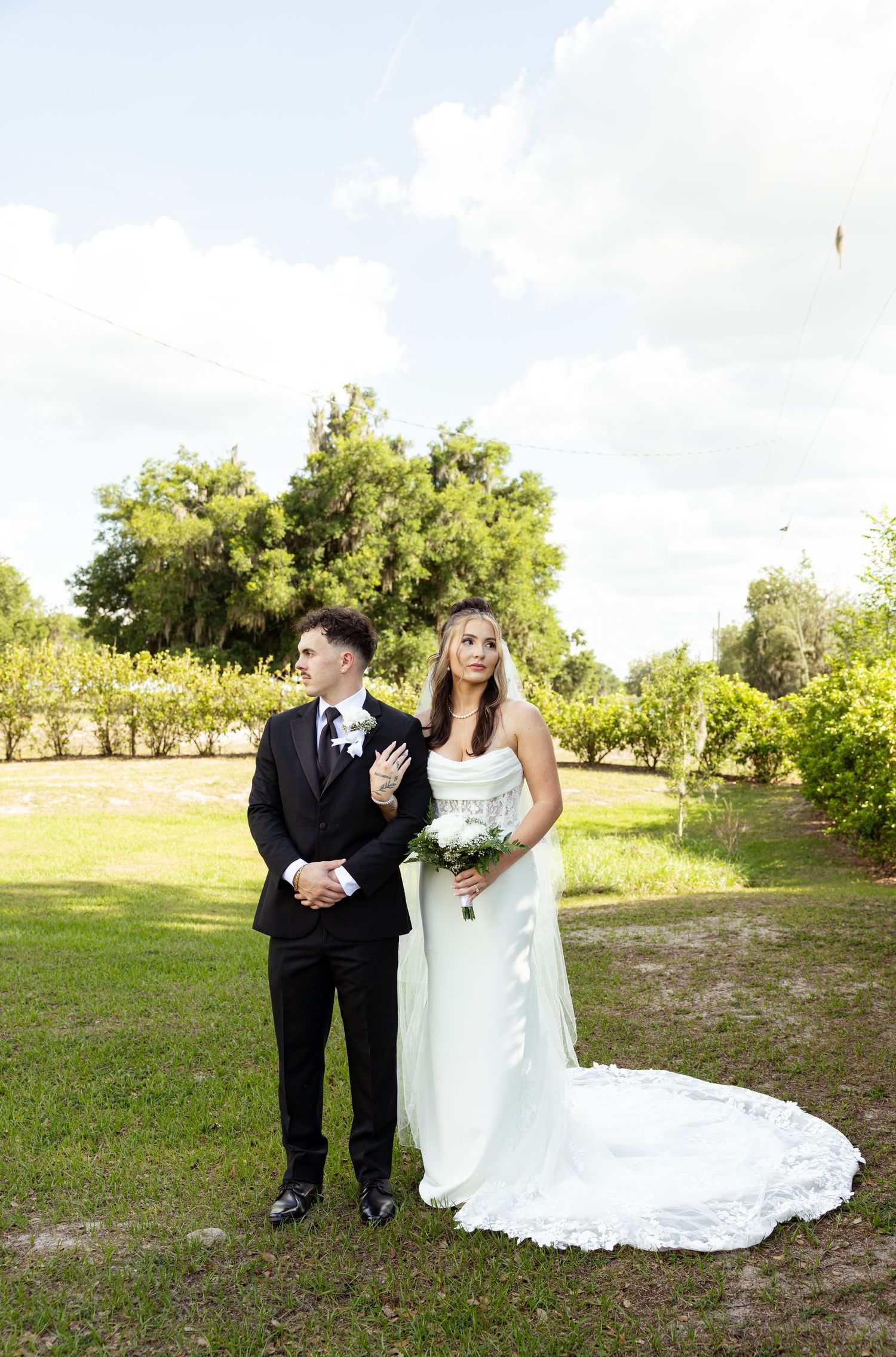 Bride in white gown holding bouquet stands with groom in black tux on green lawn with trees and cloudy sky.