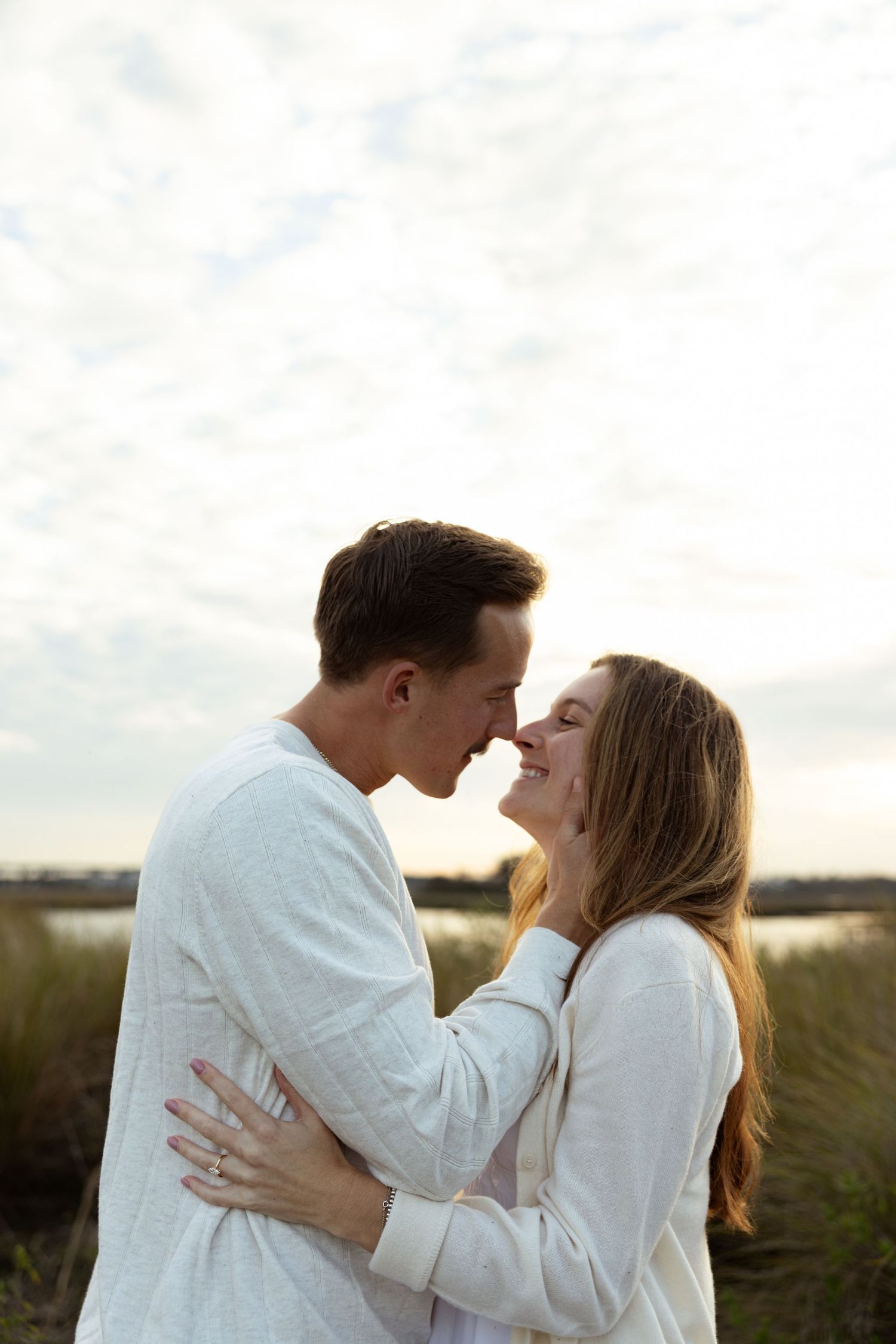 A happy couple sharing a romantic nose touch embrace outdoors near a lake at sunset, both wearing white tops.