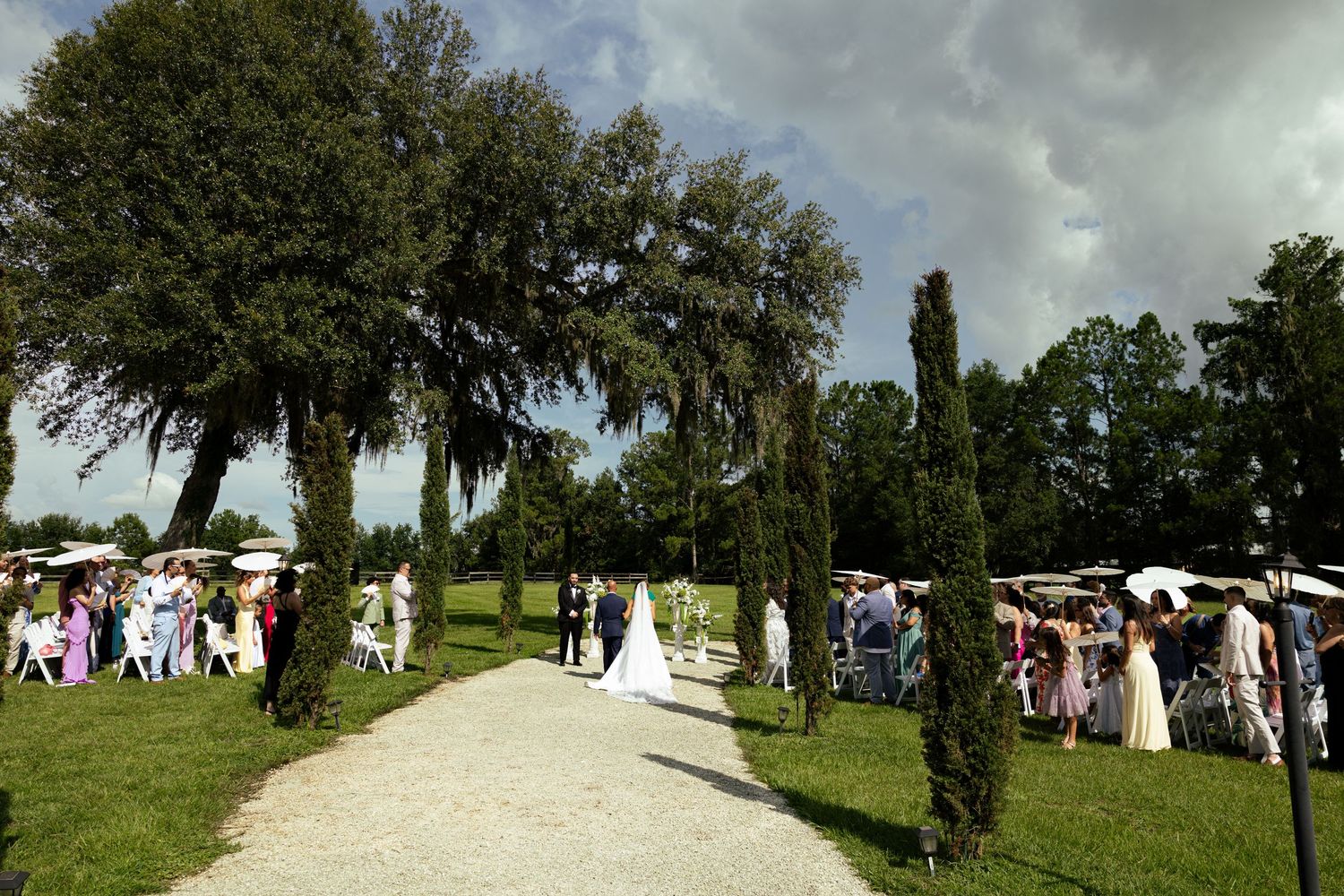 Outdoor wedding ceremony on a gravel path surrounded by tall trees and lush greenery under a dramatic cloudy sky.