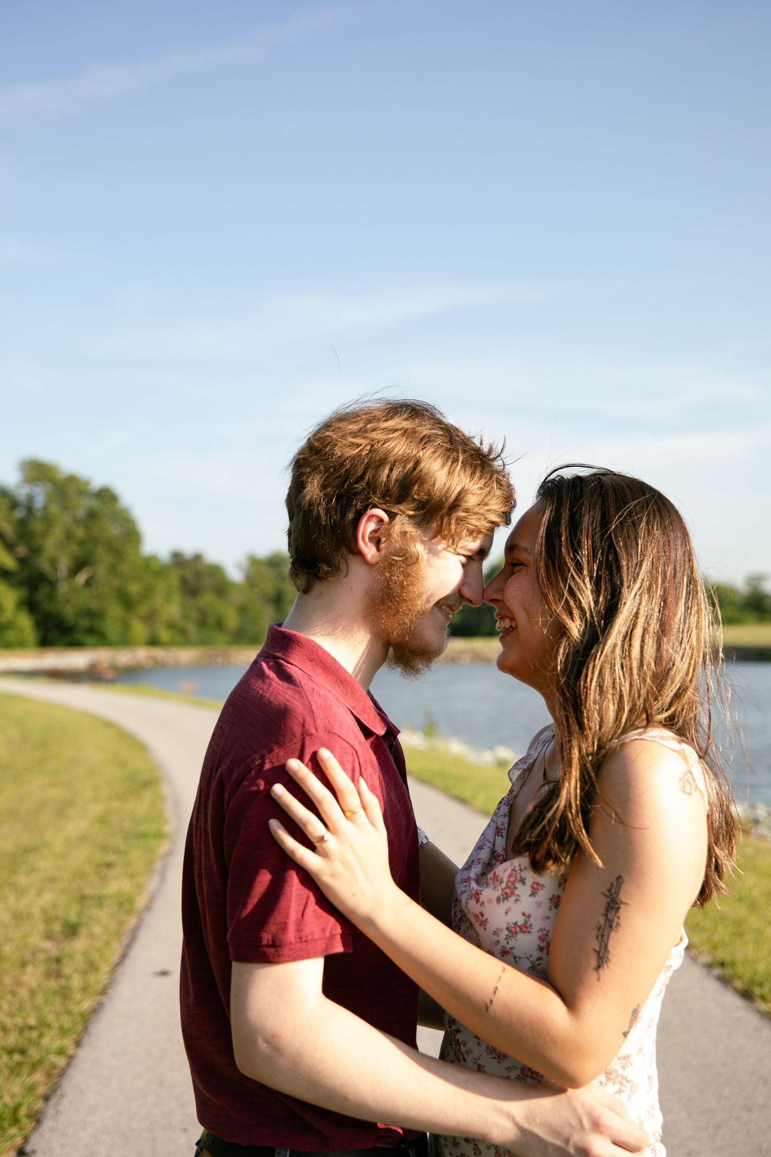 A young couple sharing a romantic moment outdoors by a lake, smiling and embracing on a sunny day.