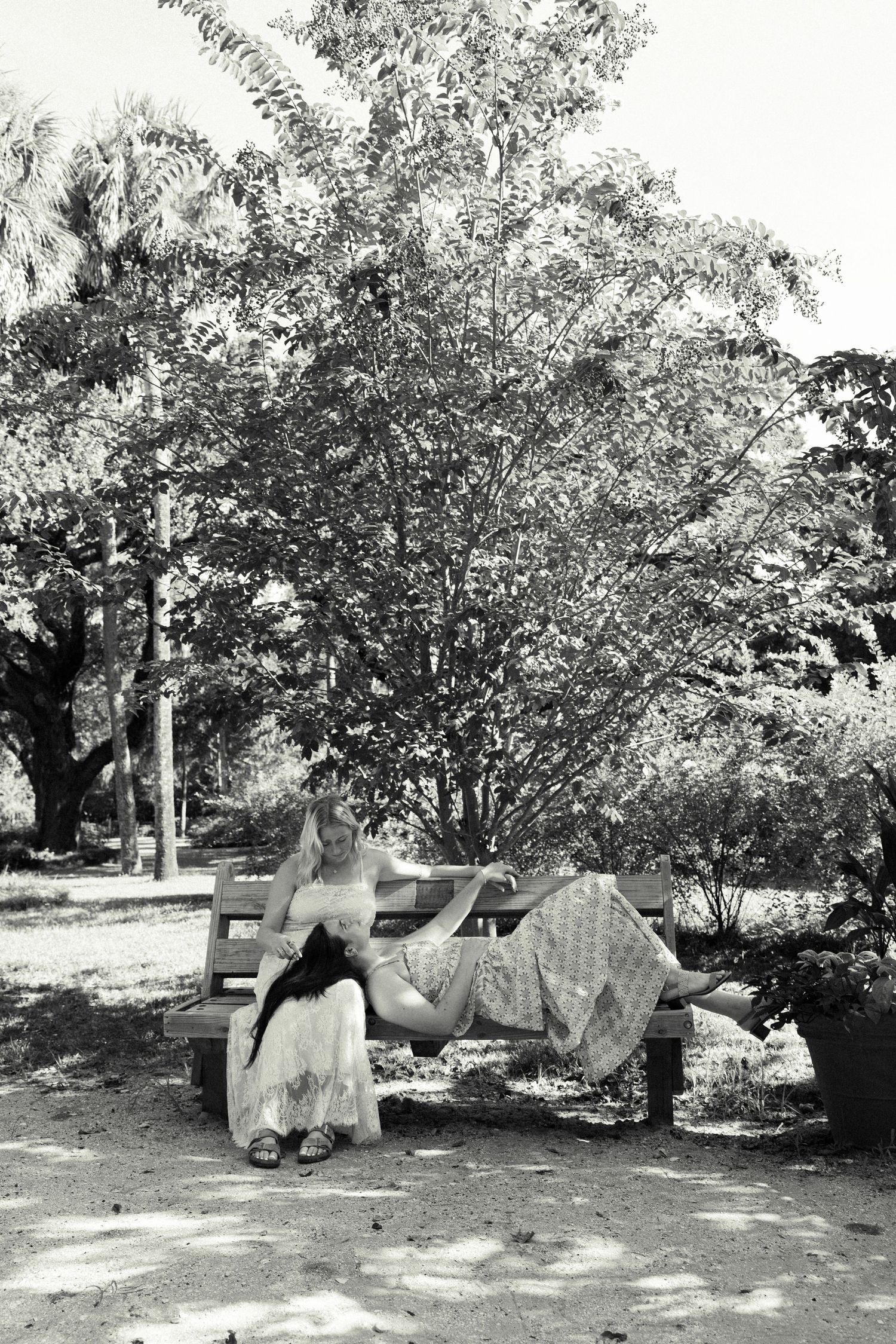 Black and white photo of two women relaxing together on a wooden park bench under lush trees.