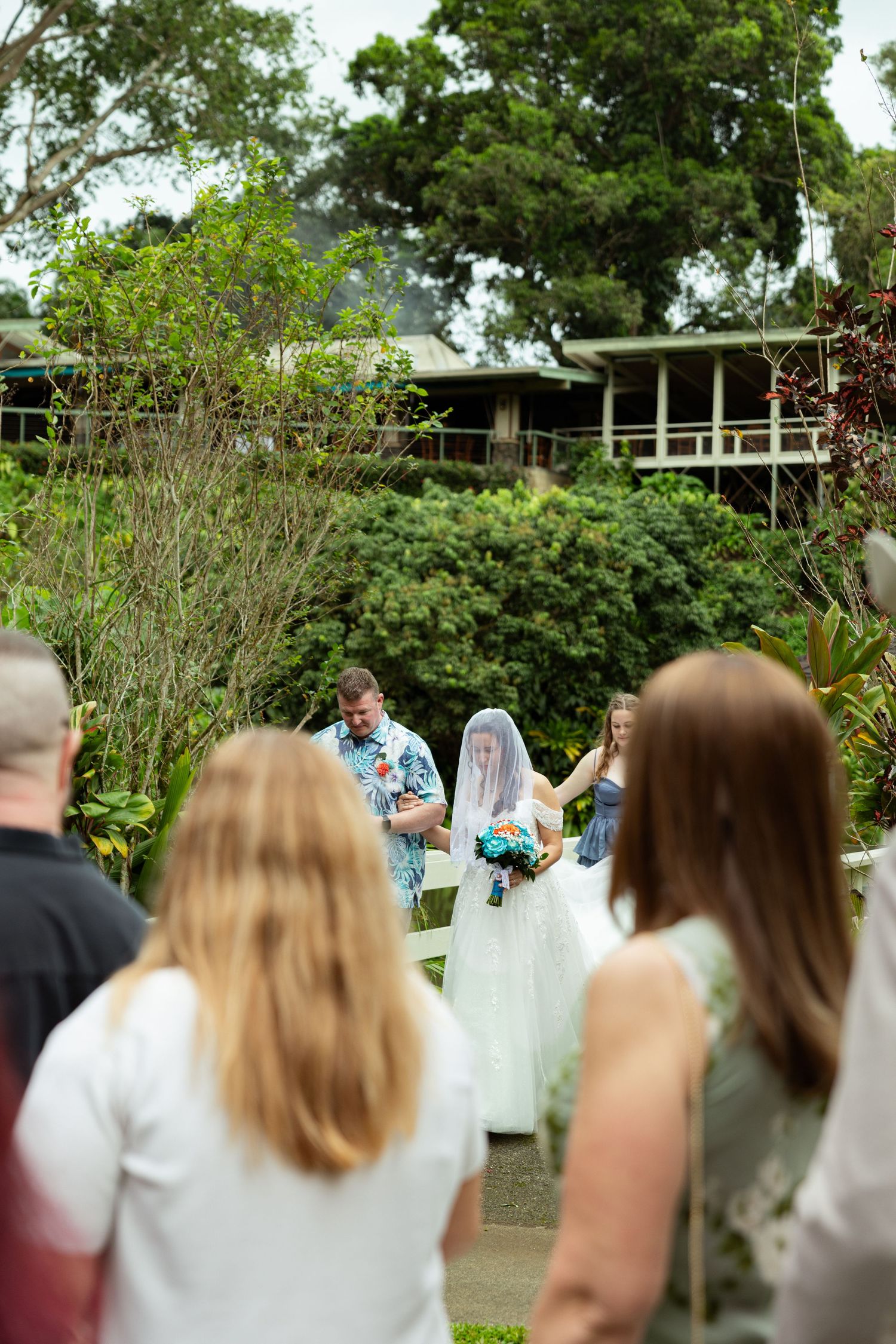 Outdoor garden wedding ceremony with bride in white gown and veil holding bouquet, guests watching near lush tropical greenery.