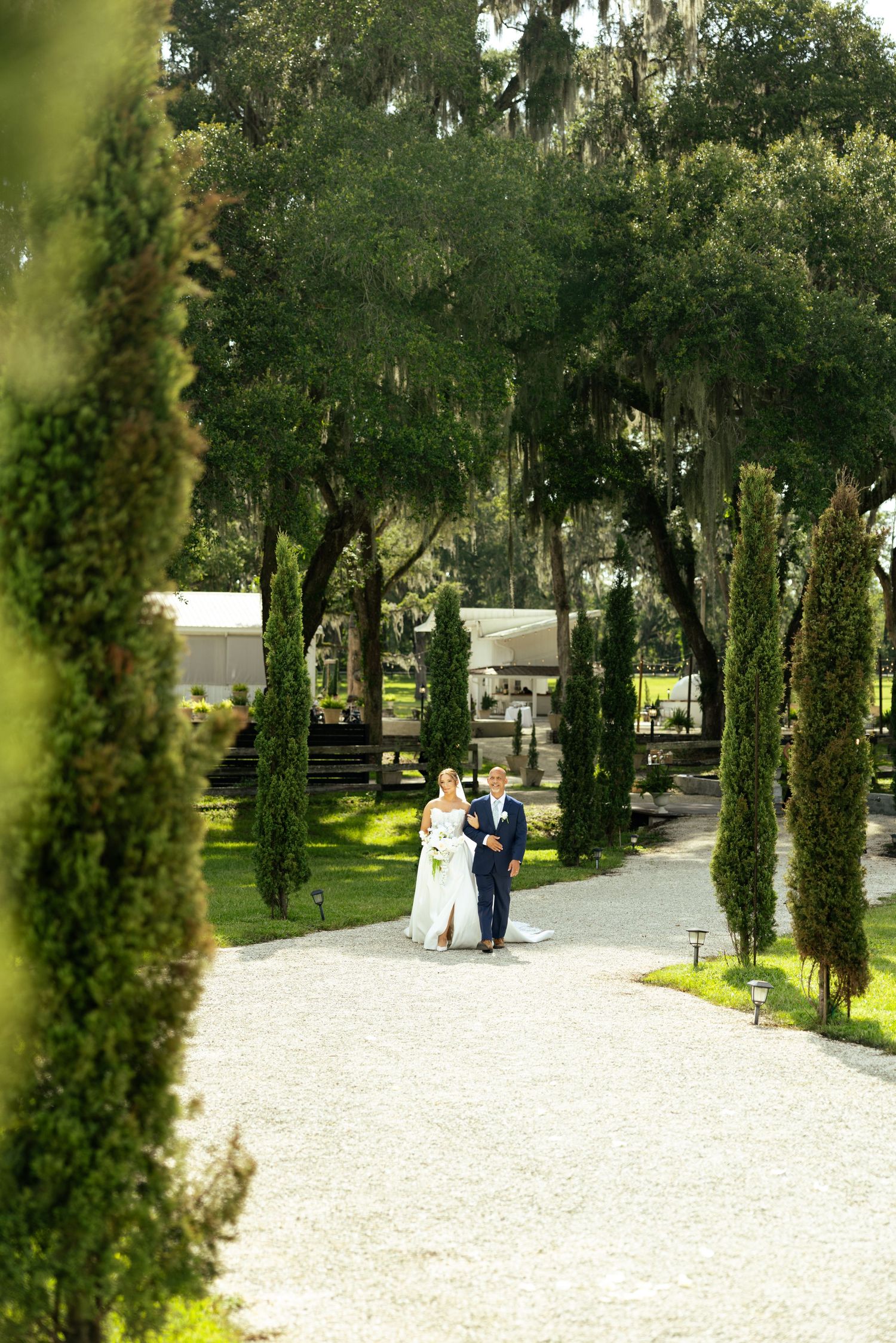 A bride and groom walk hand in hand down a tree-lined gravel path at an elegant outdoor wedding venue.