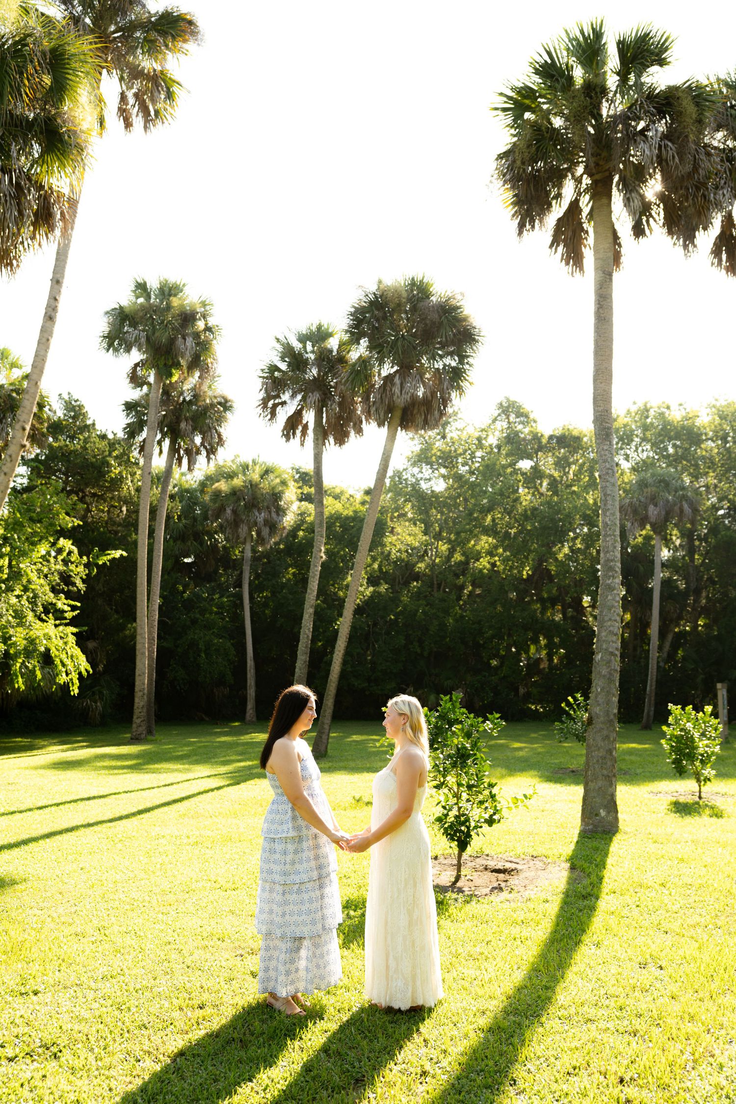 Two women in elegant dresses hold hands on a sunny green lawn surrounded by tall palm trees.