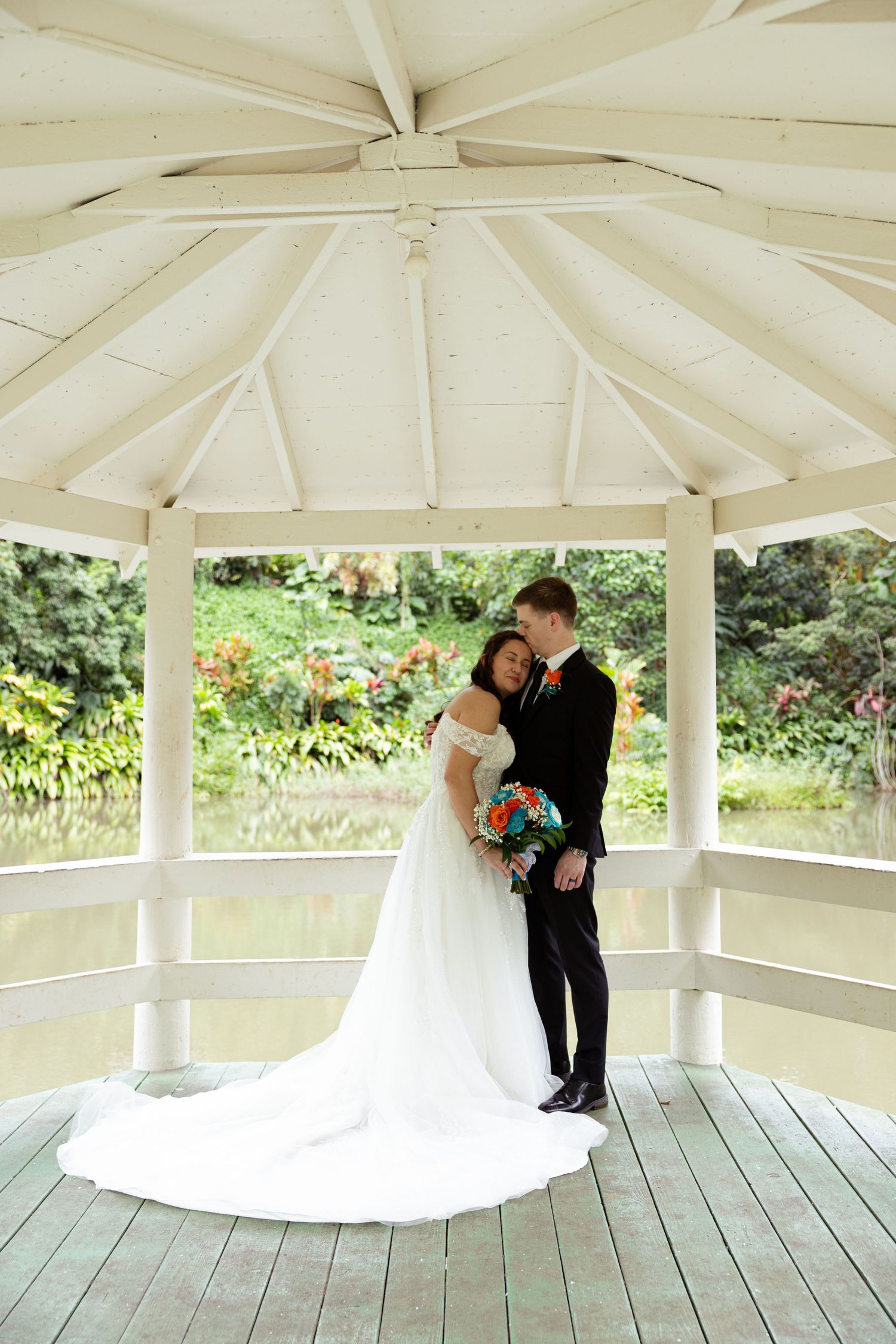 Bride and groom embrace under white gazebo with lush garden backdrop, bride holding colorful bouquet in elegant white gown.