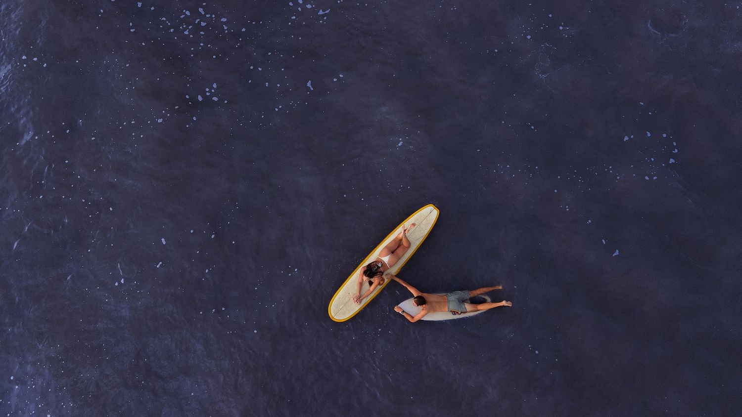 Aerial view of a surfer lying next to a yellow surfboard floating on dark ocean water.