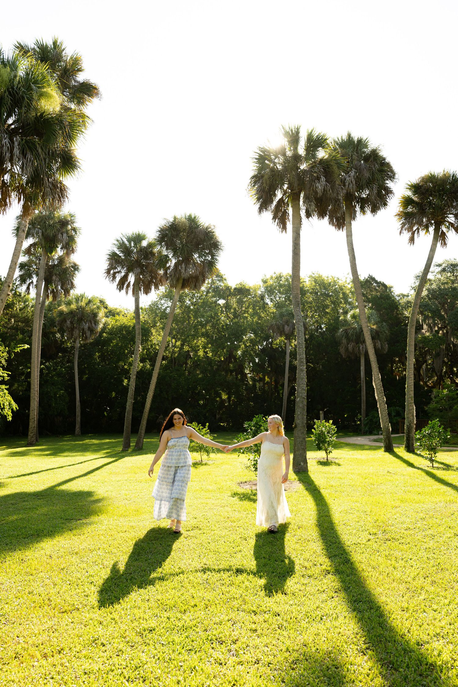 Two women in summer dresses hold hands on a sunny green lawn surrounded by tall palm trees.