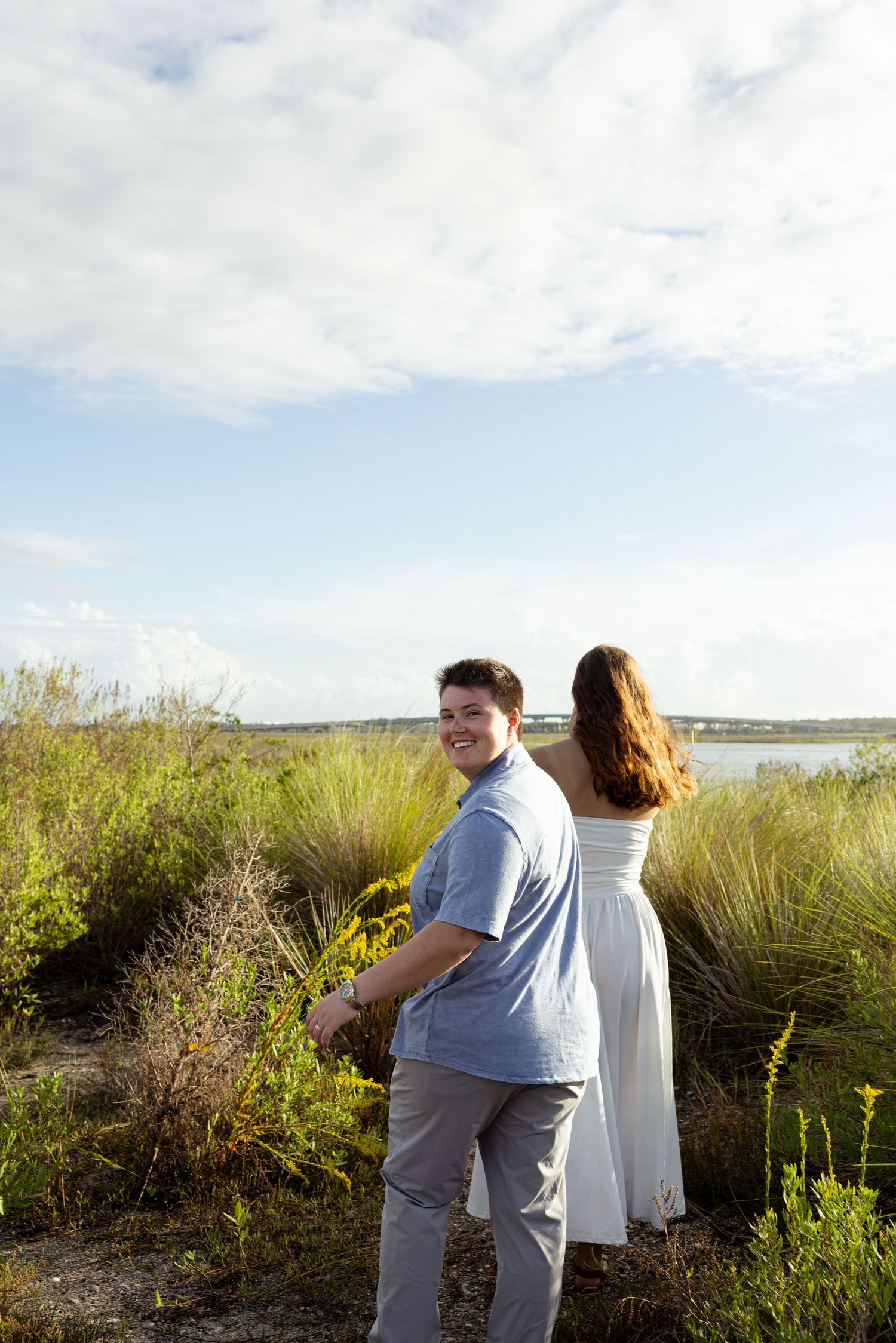 A smiling man in a blue shirt turns back while a woman in a white dress looks toward a scenic waterway.