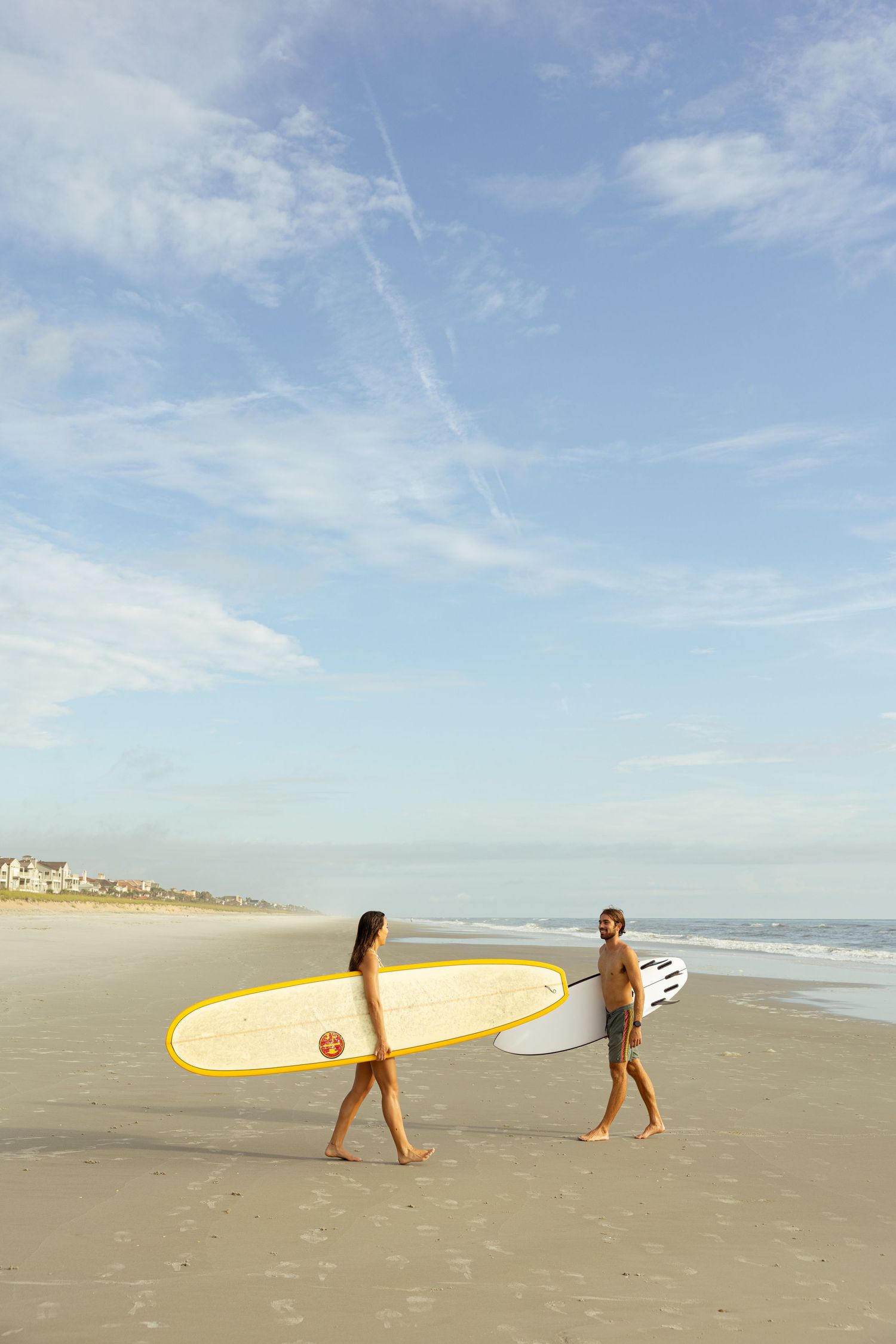 Two surfers carrying surfboards walk along a sandy beach under a partly cloudy blue sky near the ocean.