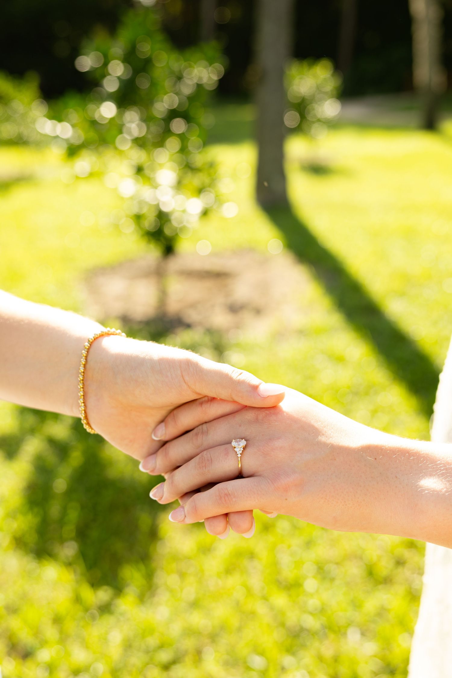 Two hands holding each other outdoors, one wearing a diamond engagement ring and gold bracelet, with green grass background.