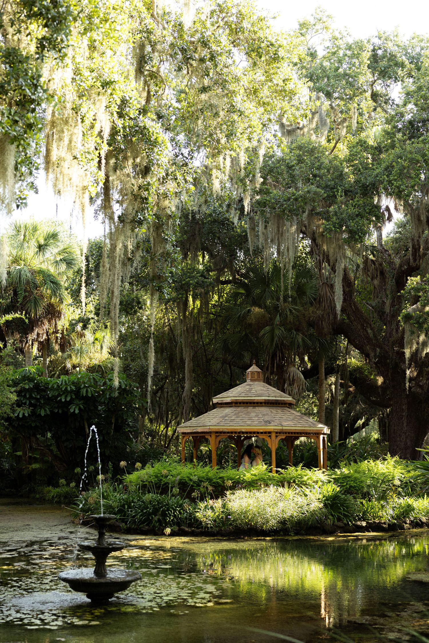 Serene garden gazebo beside a lily pond with a fountain, surrounded by lush greenery and Spanish moss-draped oak trees.