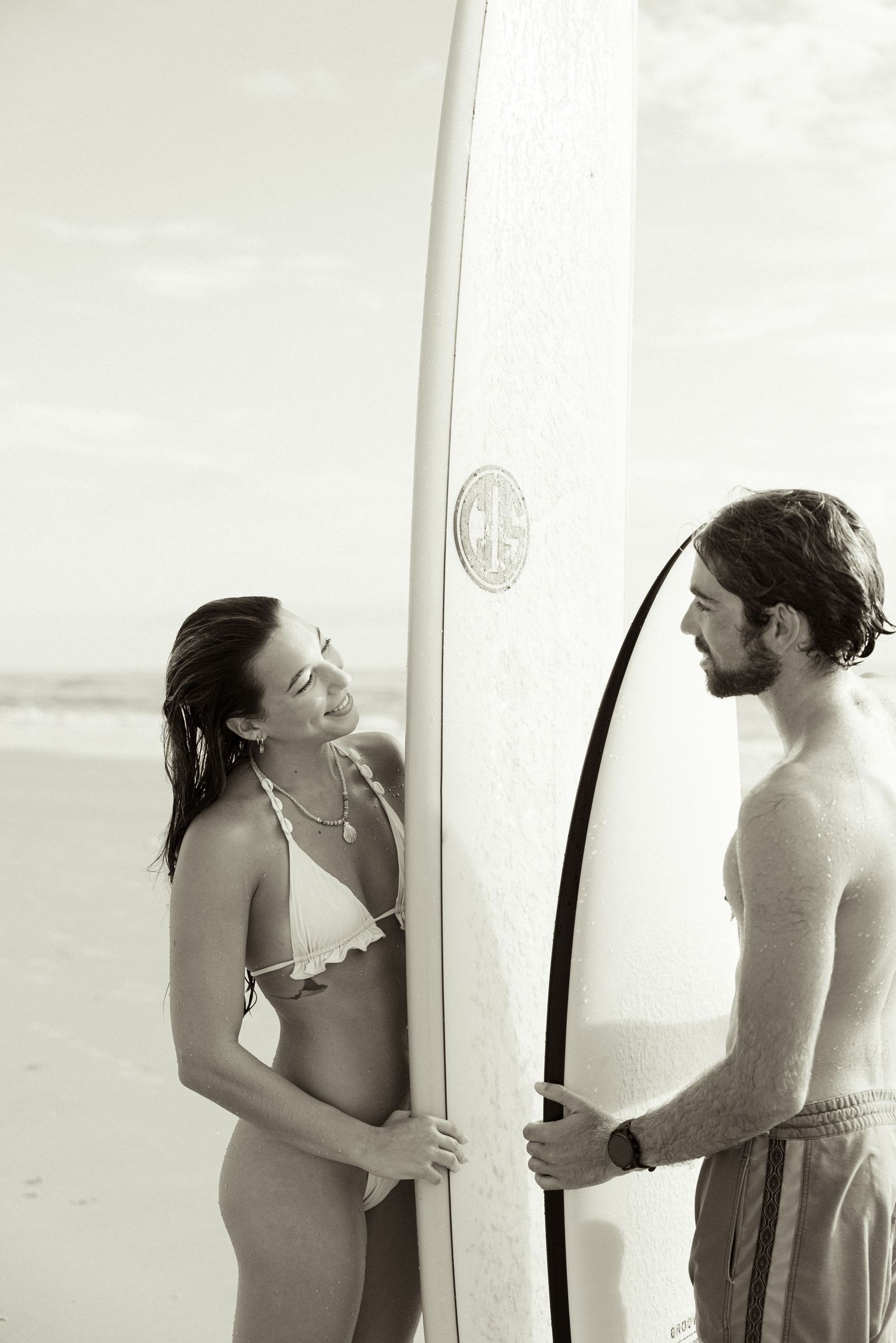 Black and white photo of a couple smiling and holding a surfboard together on a sandy beach.