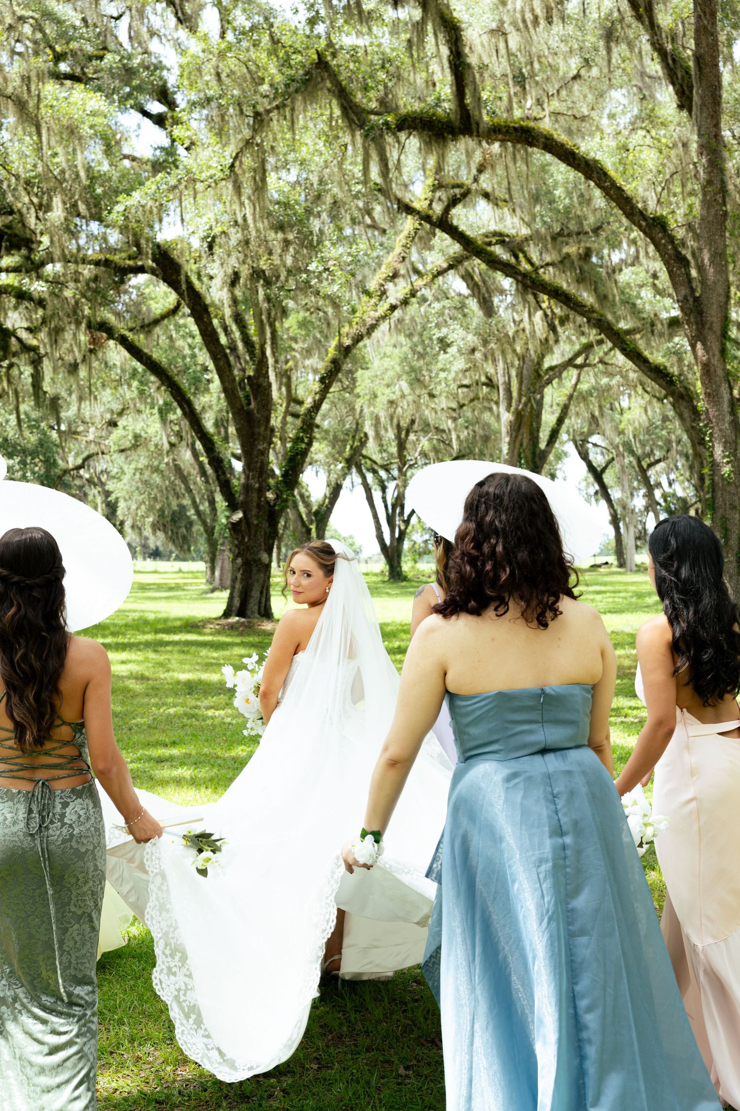 Bride walking with bridesmaids in blue and pink dresses under moss-draped oak trees at an outdoor wedding.