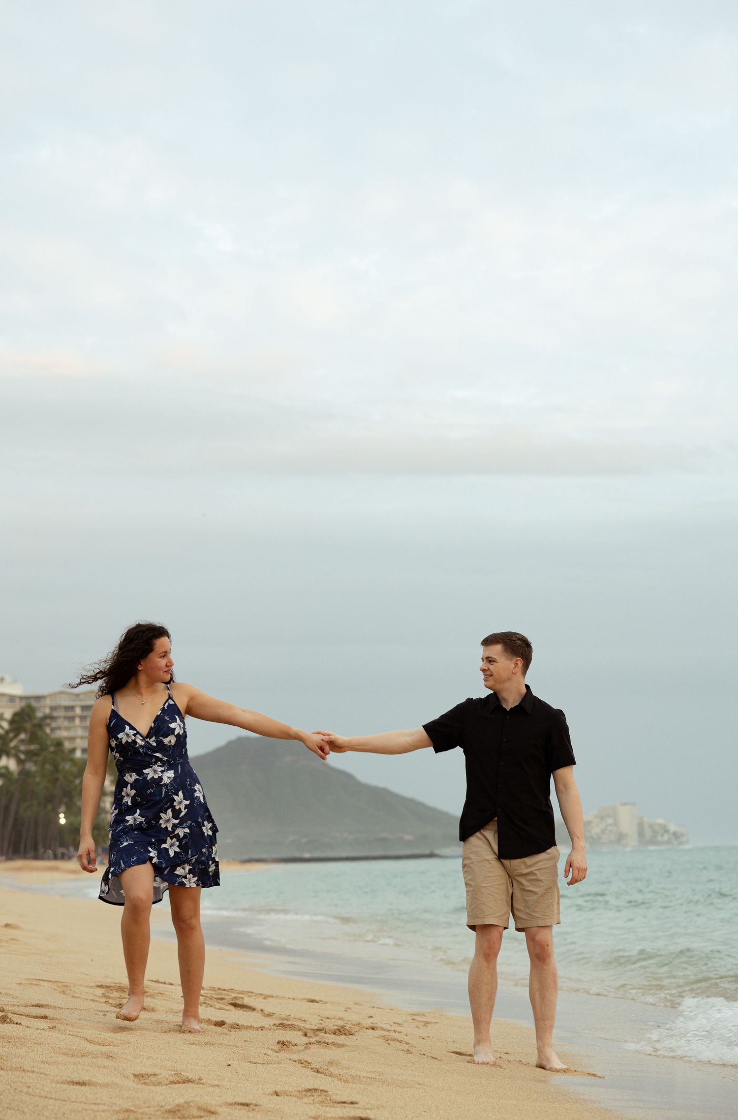 Couple holding hands and walking barefoot on a sandy beach with ocean waves and mountains in the background.