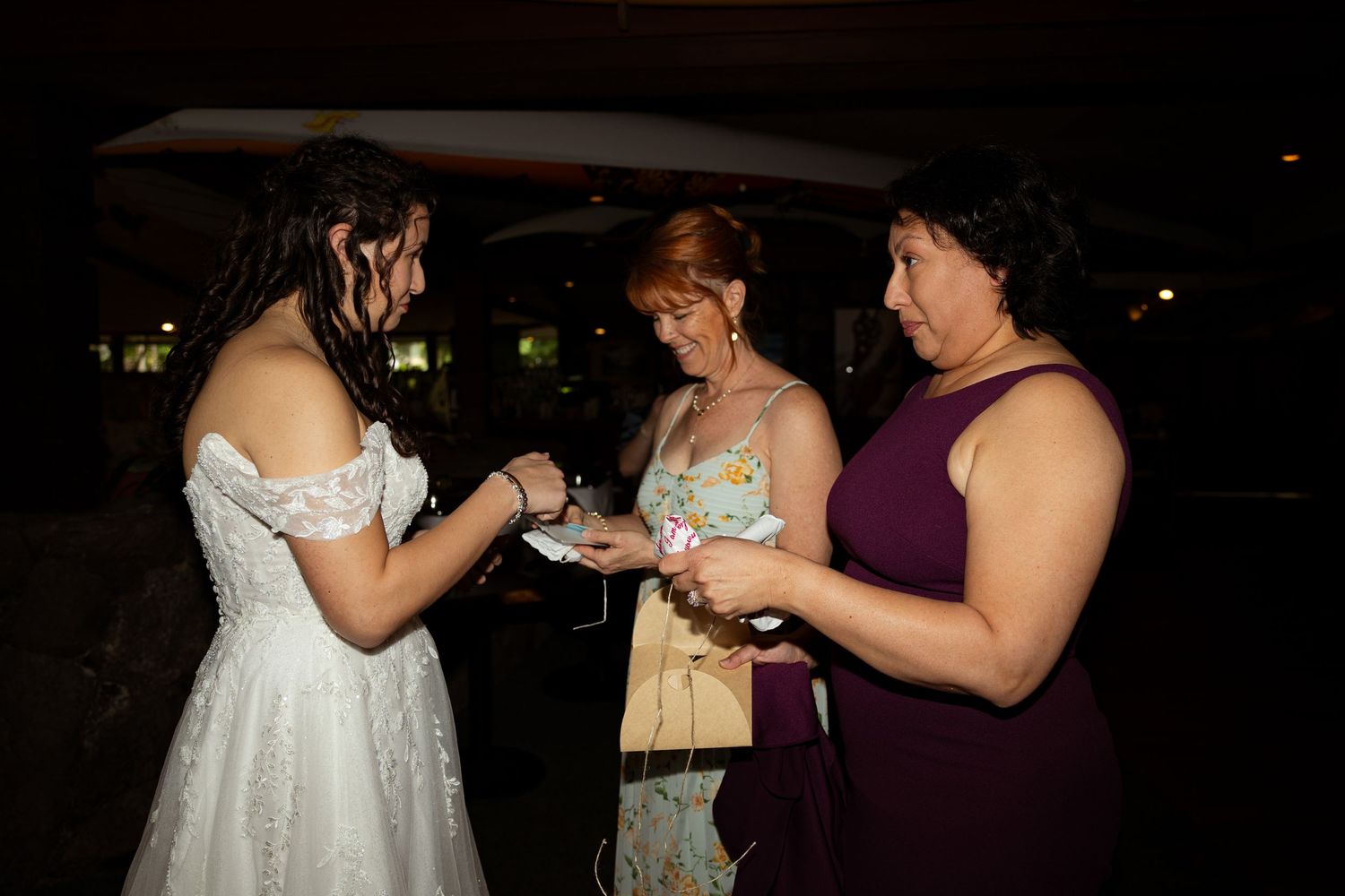 Bride in white dress exchanging items with two women in formal attire at a dark evening wedding reception.