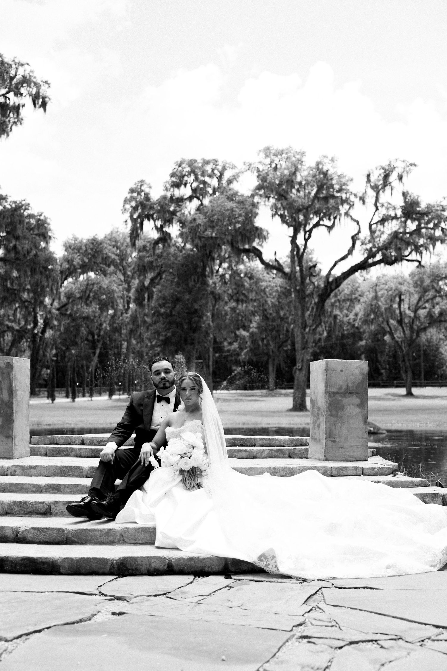 Black and white wedding photo of bride and groom sitting on stone steps surrounded by moss-draped trees.