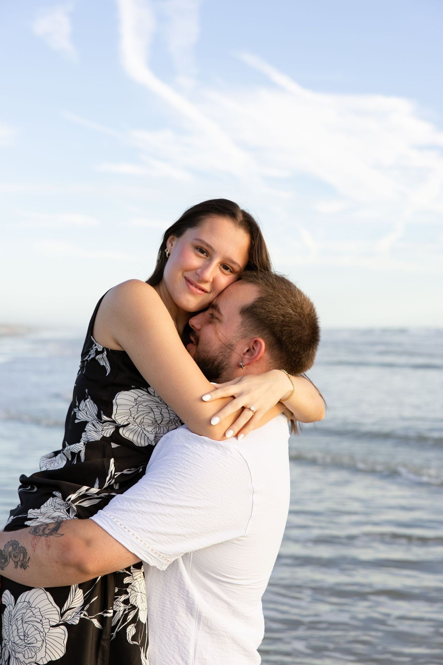 Happy woman in floral dress embracing her partner on the beach with blue sky and ocean in the background.